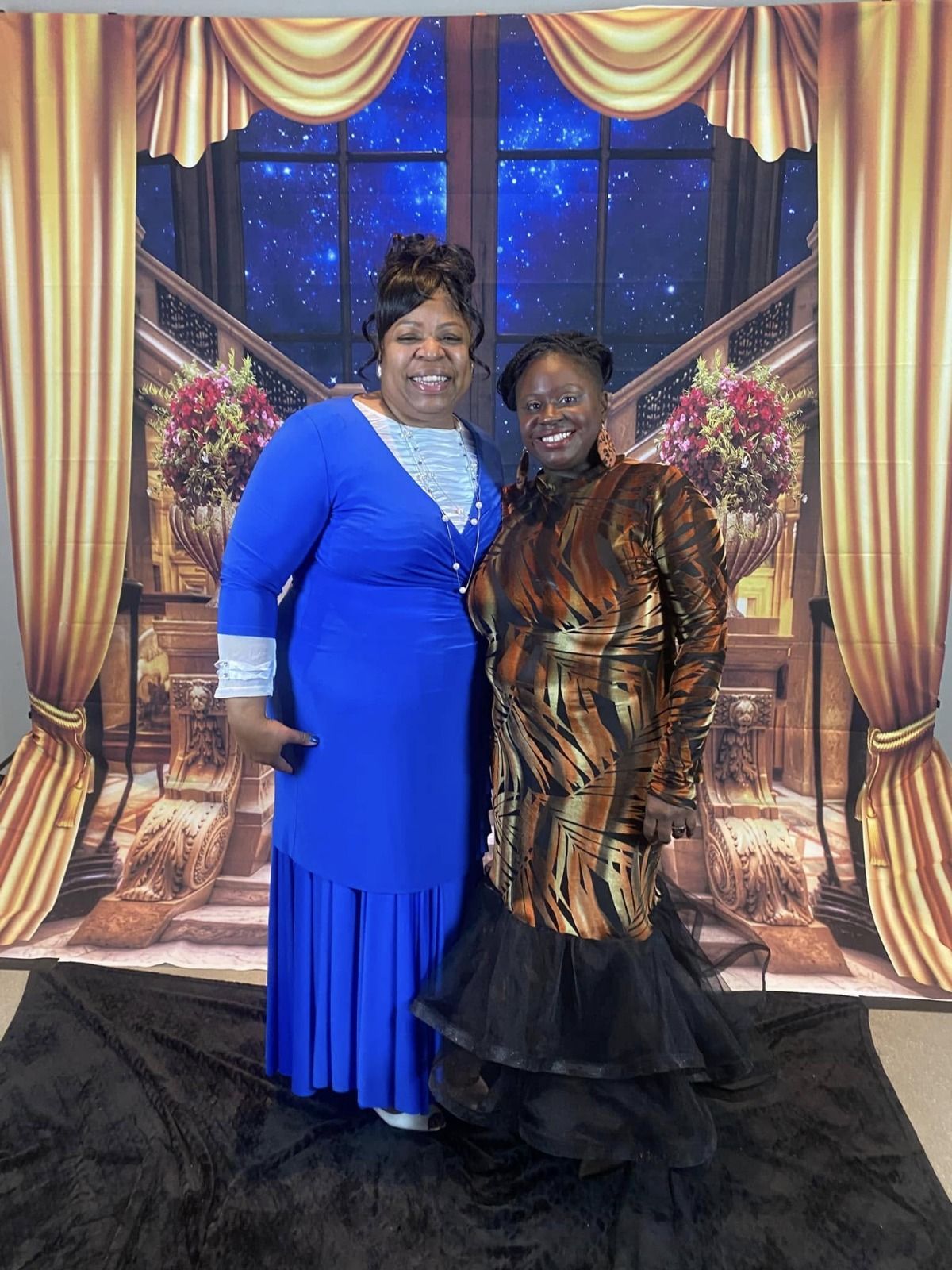 Two women pose in front of a backdrop, one in blue dress, the other in a patterned top and tiered black skirt.