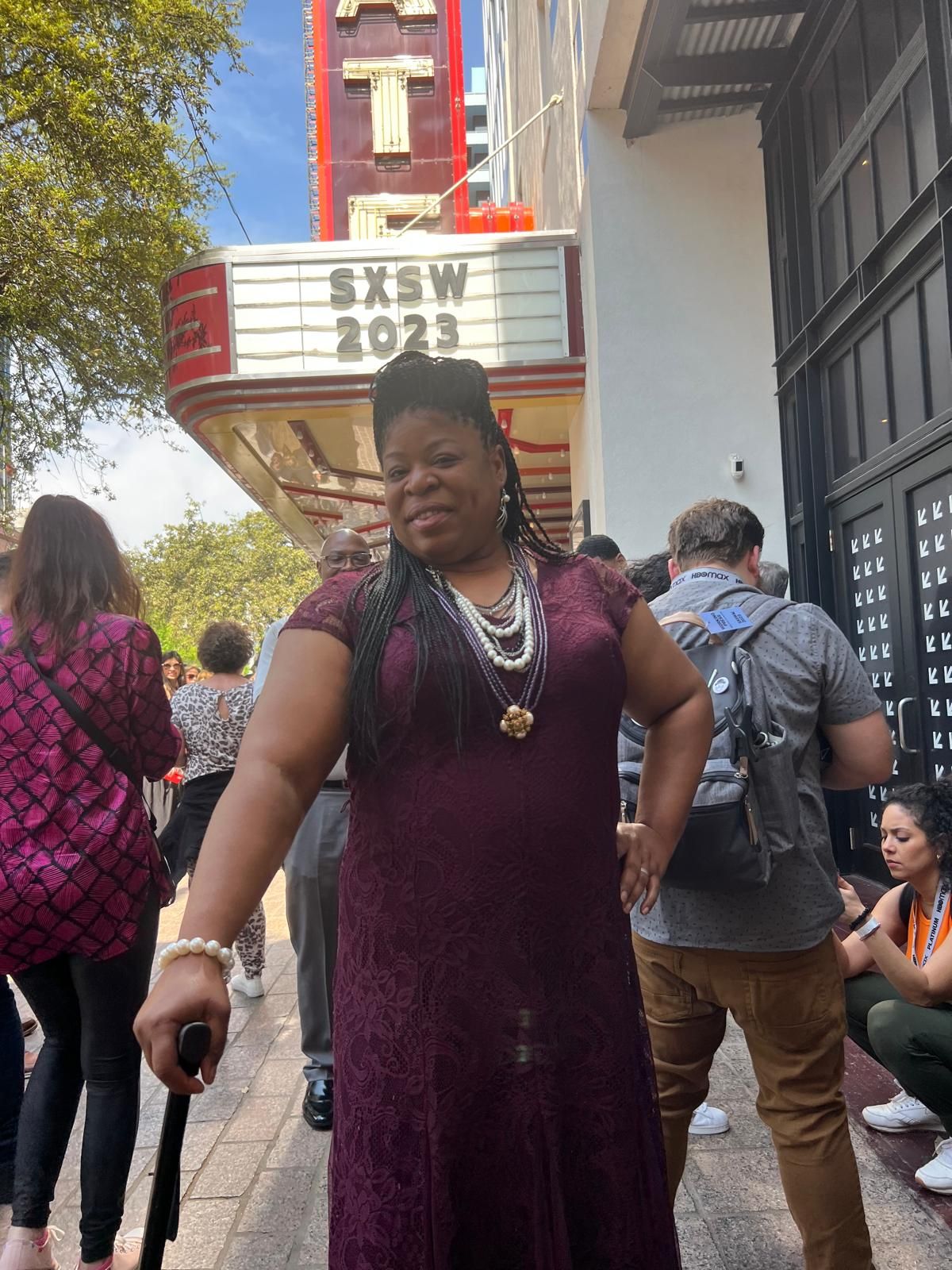 Woman in a purple dress poses in front of the SXSW 2023 marquee. She holds a cane and smiles.