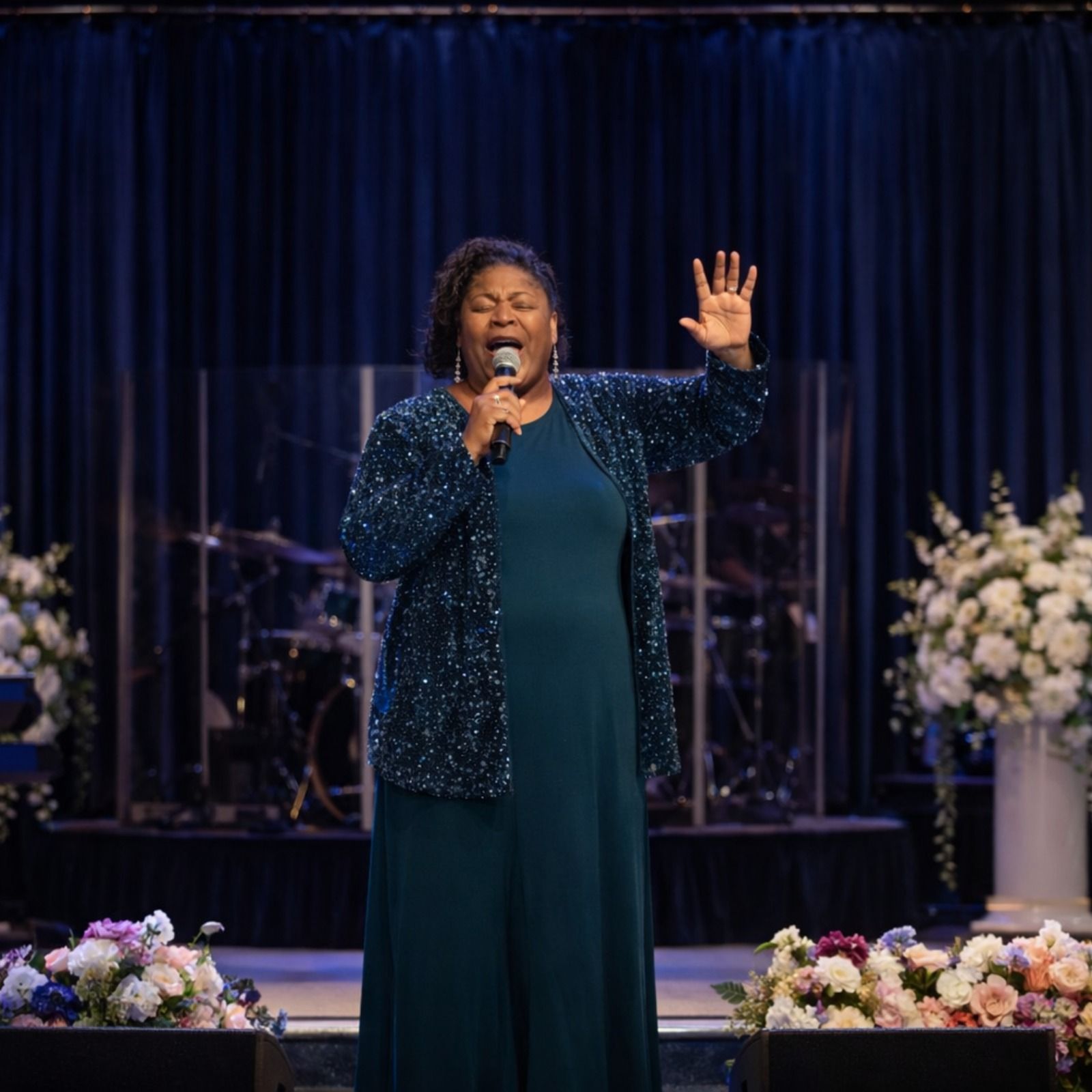 Woman speaking at a church, holding a microphone, with a raised hand. The backdrop has a banner that reads