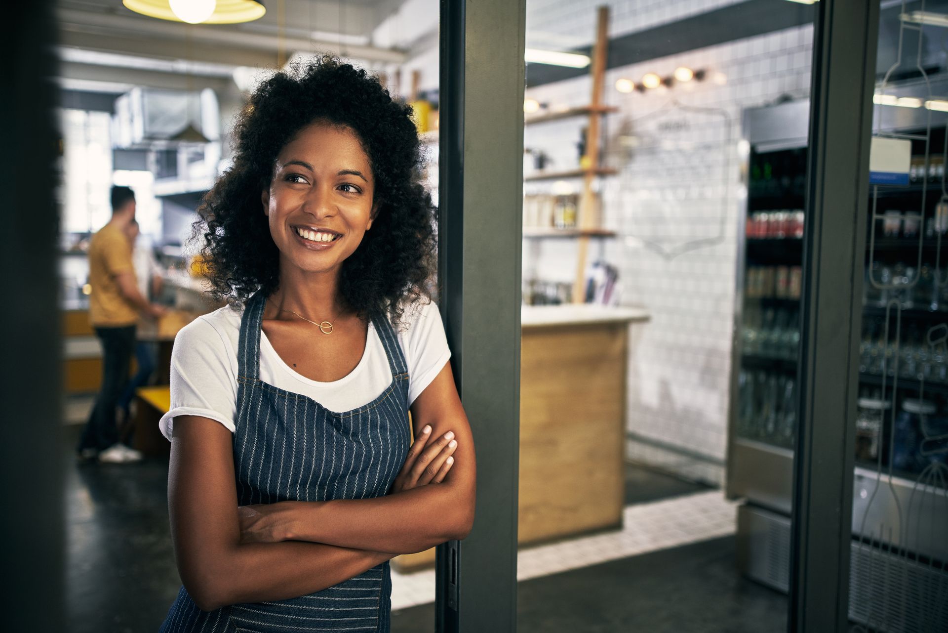Smiling Black woman in apron, arms crossed, doorway of business.