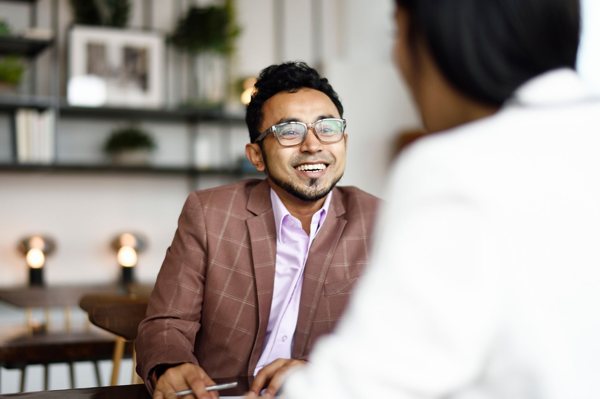 Man with glasses and brown blazer smiling, in conversation with a person in white.