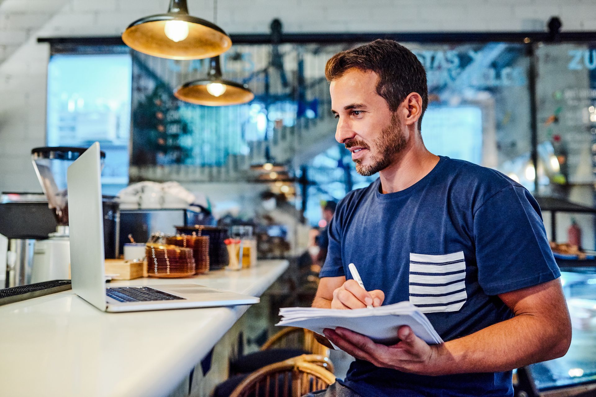 Man in cafe taking notes while looking at a laptop on counter.