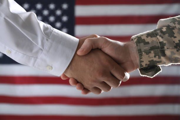 Two people shaking hands in front of an American flag. One wears a white shirt, the other military camouflage.