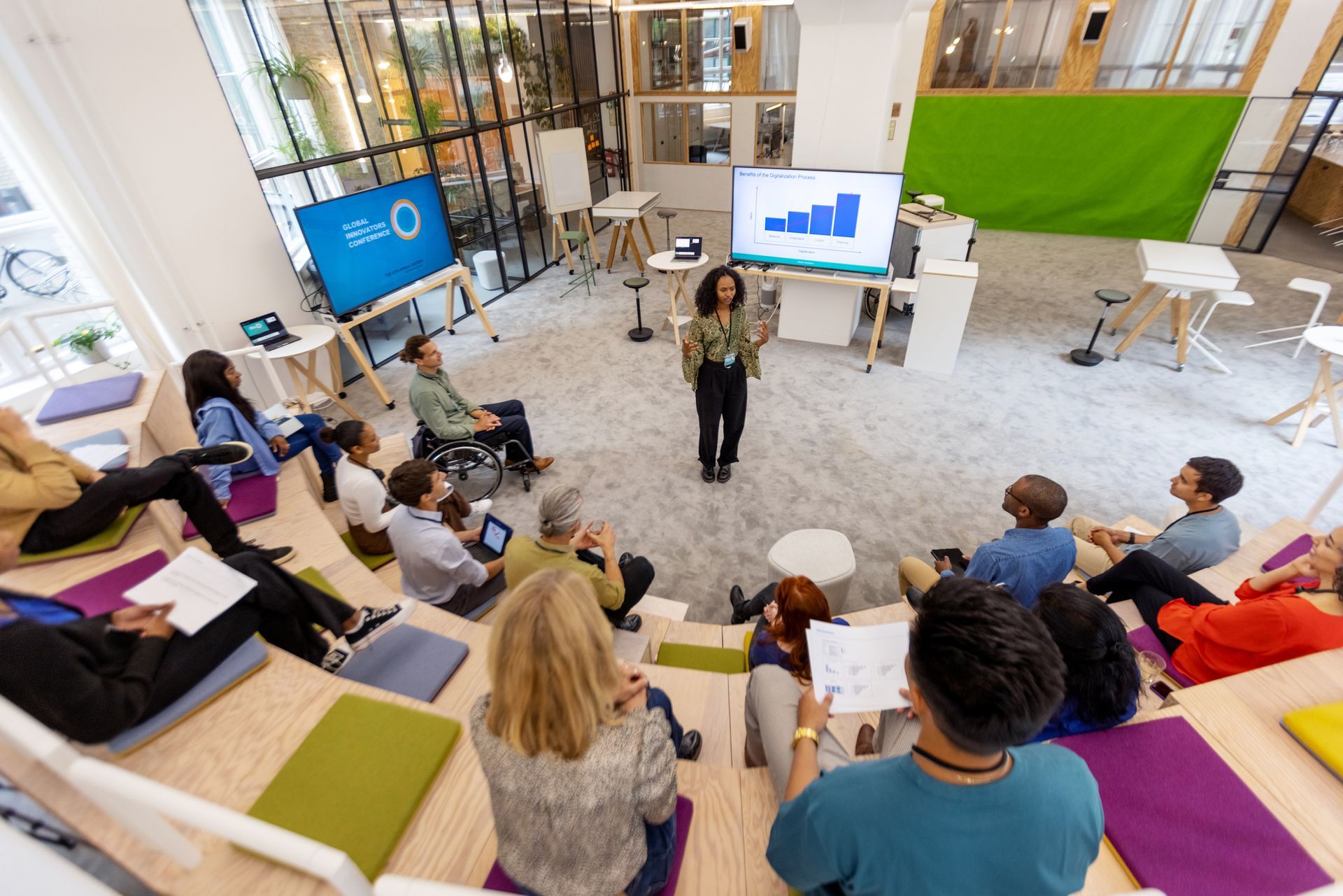 Woman presenting data to diverse group in modern office, tiered seating, screens with charts, natural light.