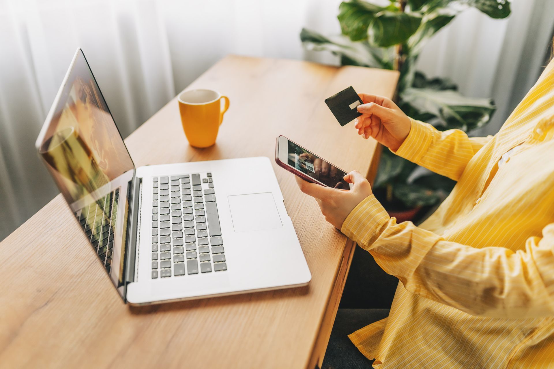 Person in yellow shirt making an online purchase with laptop, phone, and credit card.