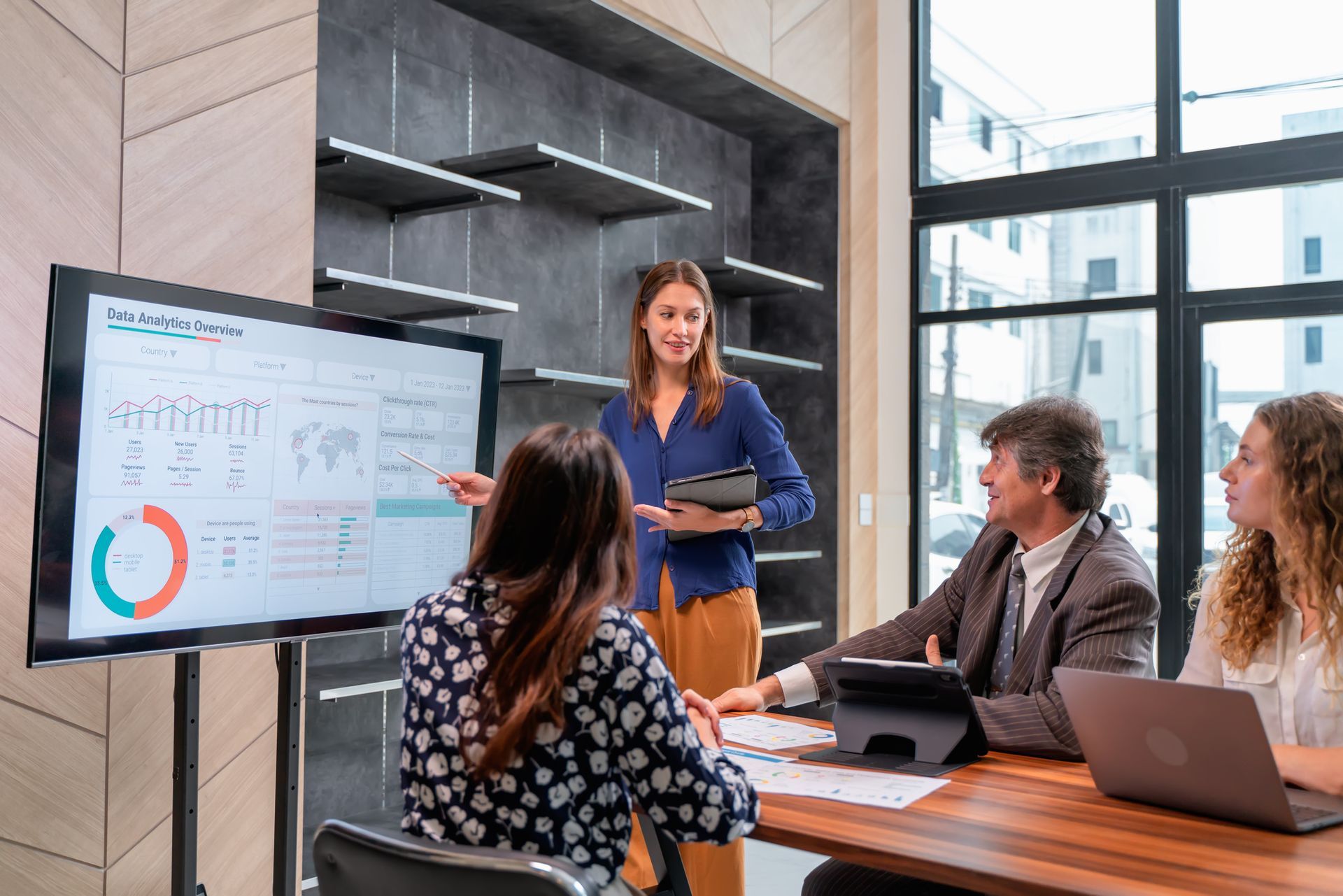 A woman points at a screen with data, presenting to a group in a modern office.