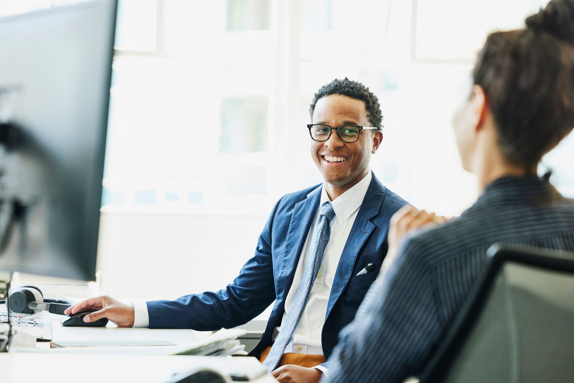 Smiling Black man in a suit at a desk with a coworker, looking at the camera in a bright office.