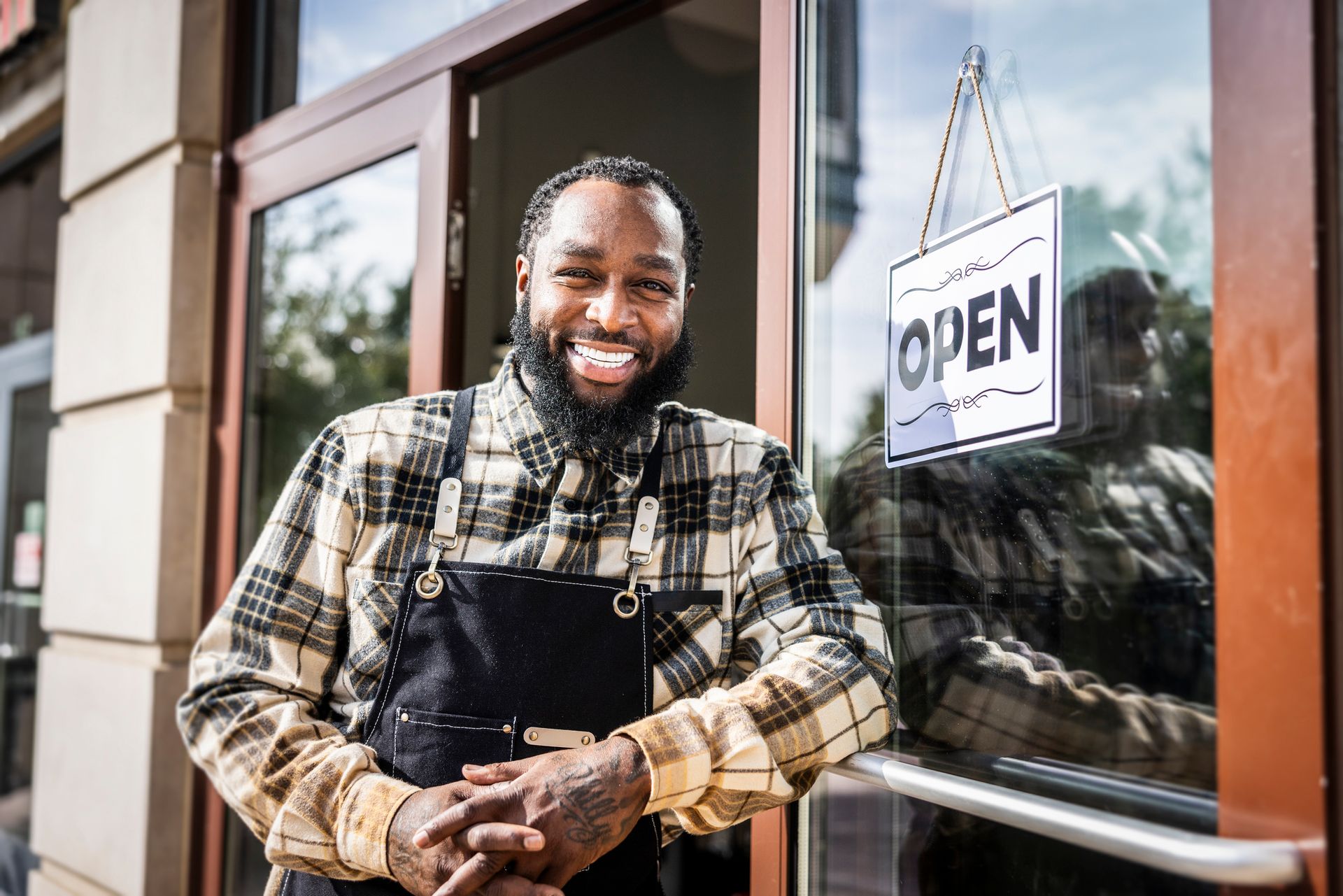 Smiling Black man in apron stands by open shop door.