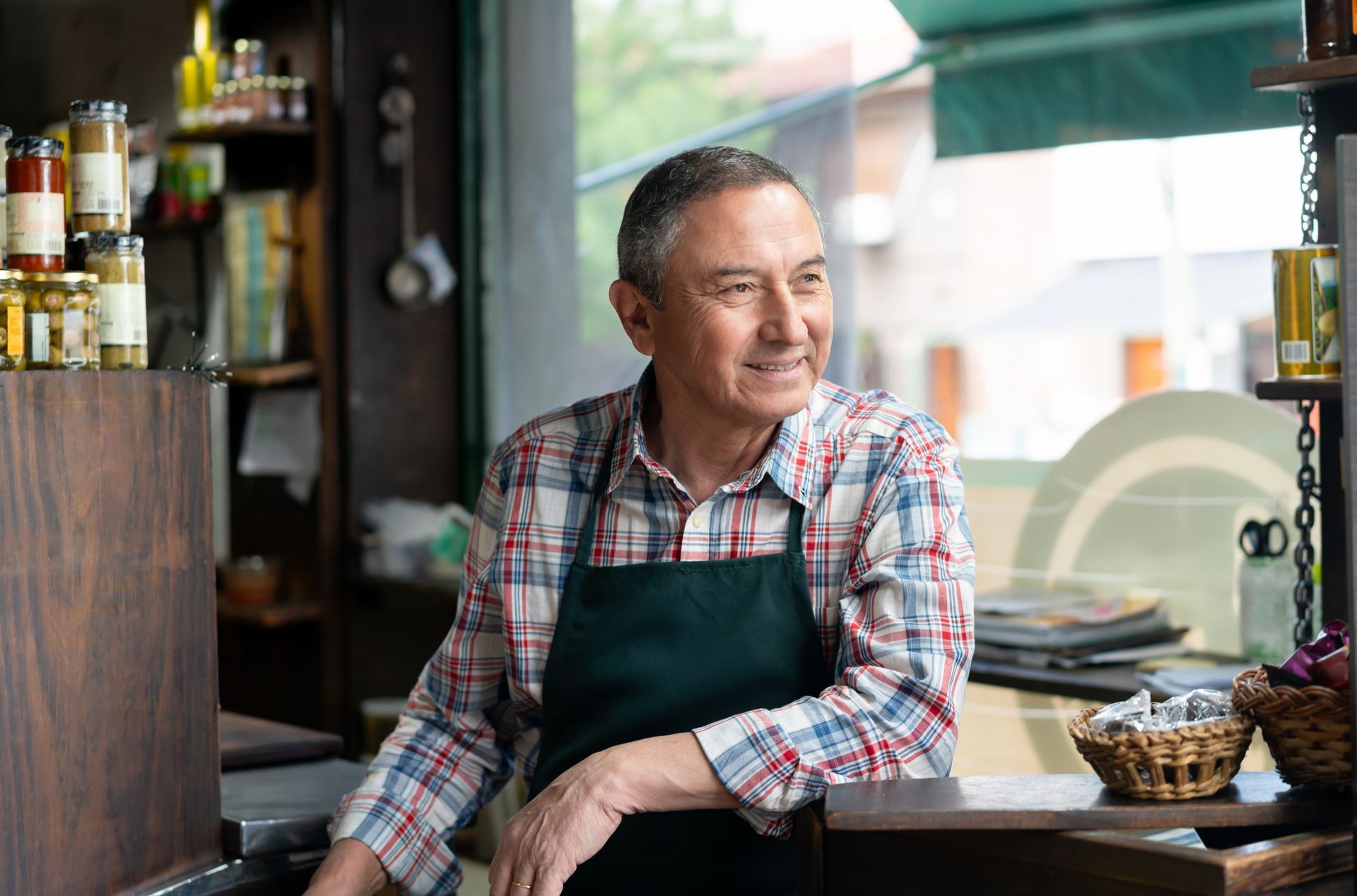 Man in apron smiles while leaning on counter of a small shop.