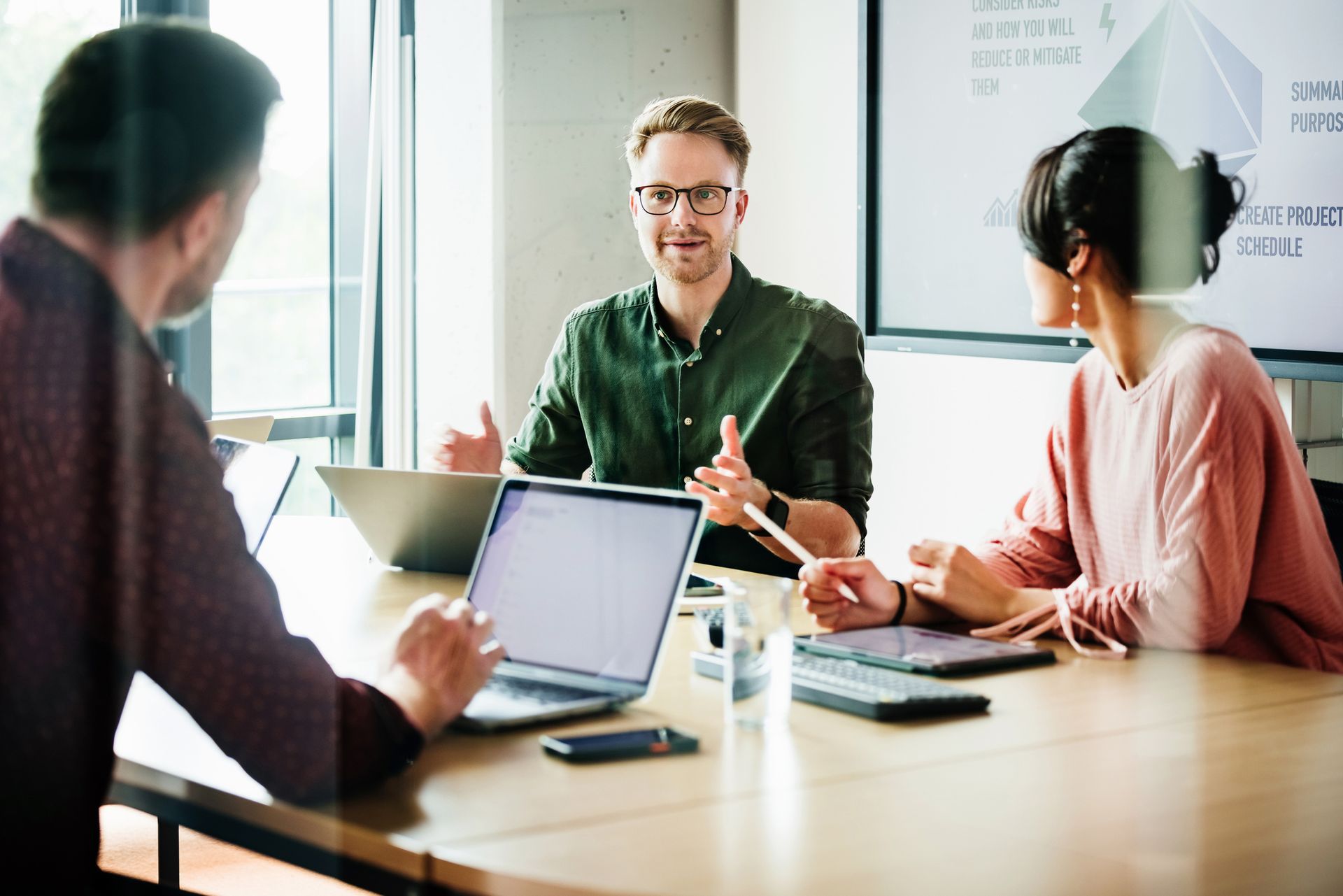 Three people in a meeting around a table with laptops, discussing a project.