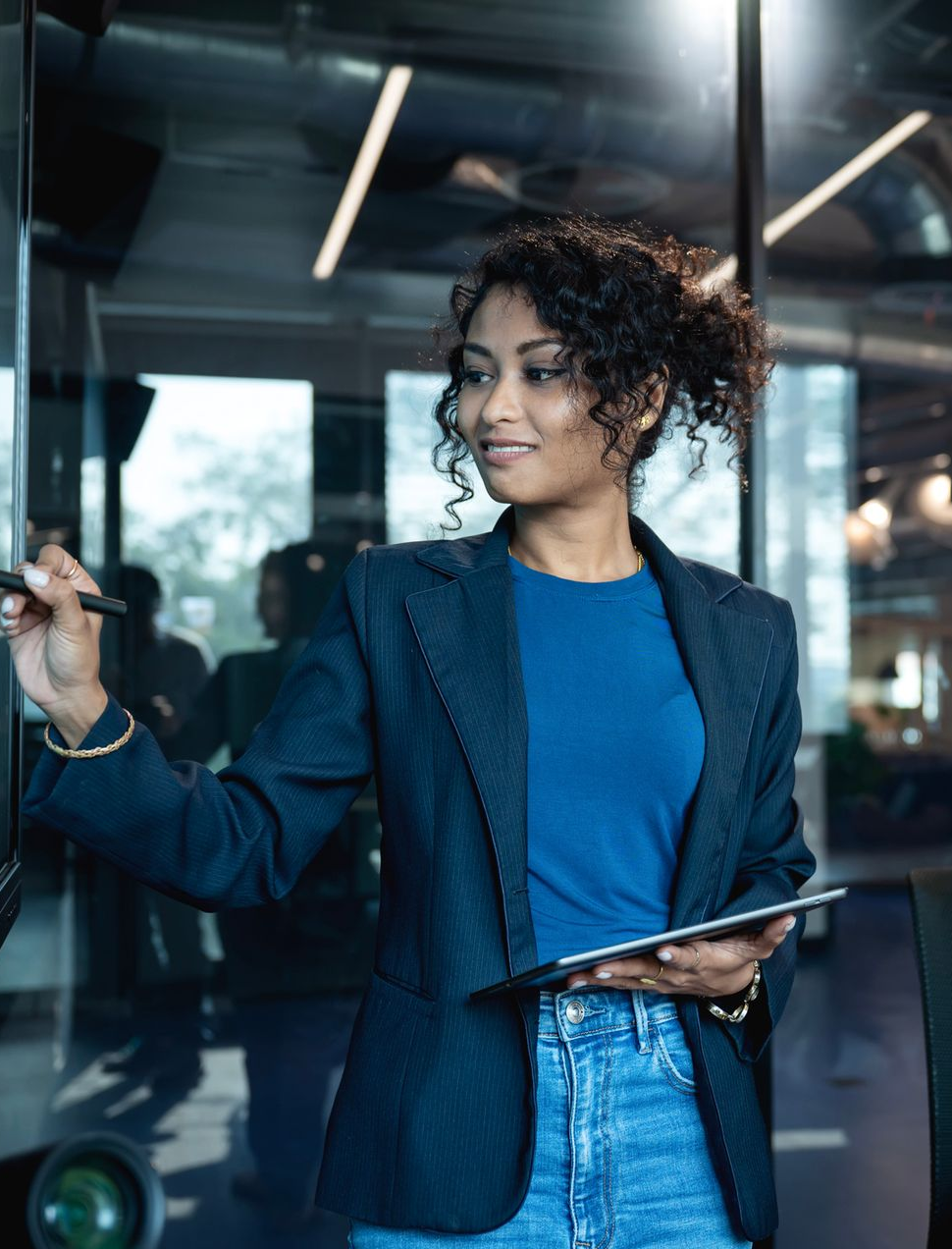 Woman in blazer writes on a whiteboard, holding a tablet, while another woman looks on in a modern office.