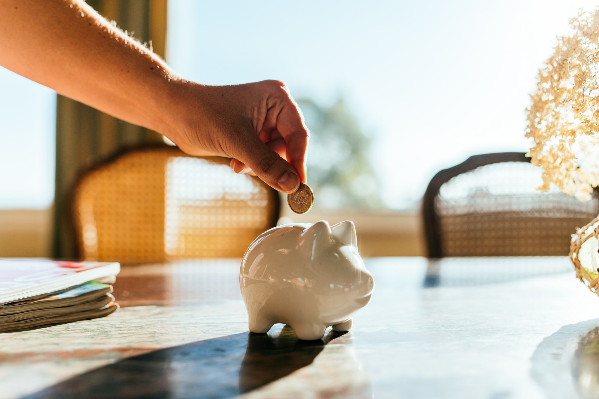 Hand dropping coin into a white piggy bank on a table. Sunlight streams through a window.