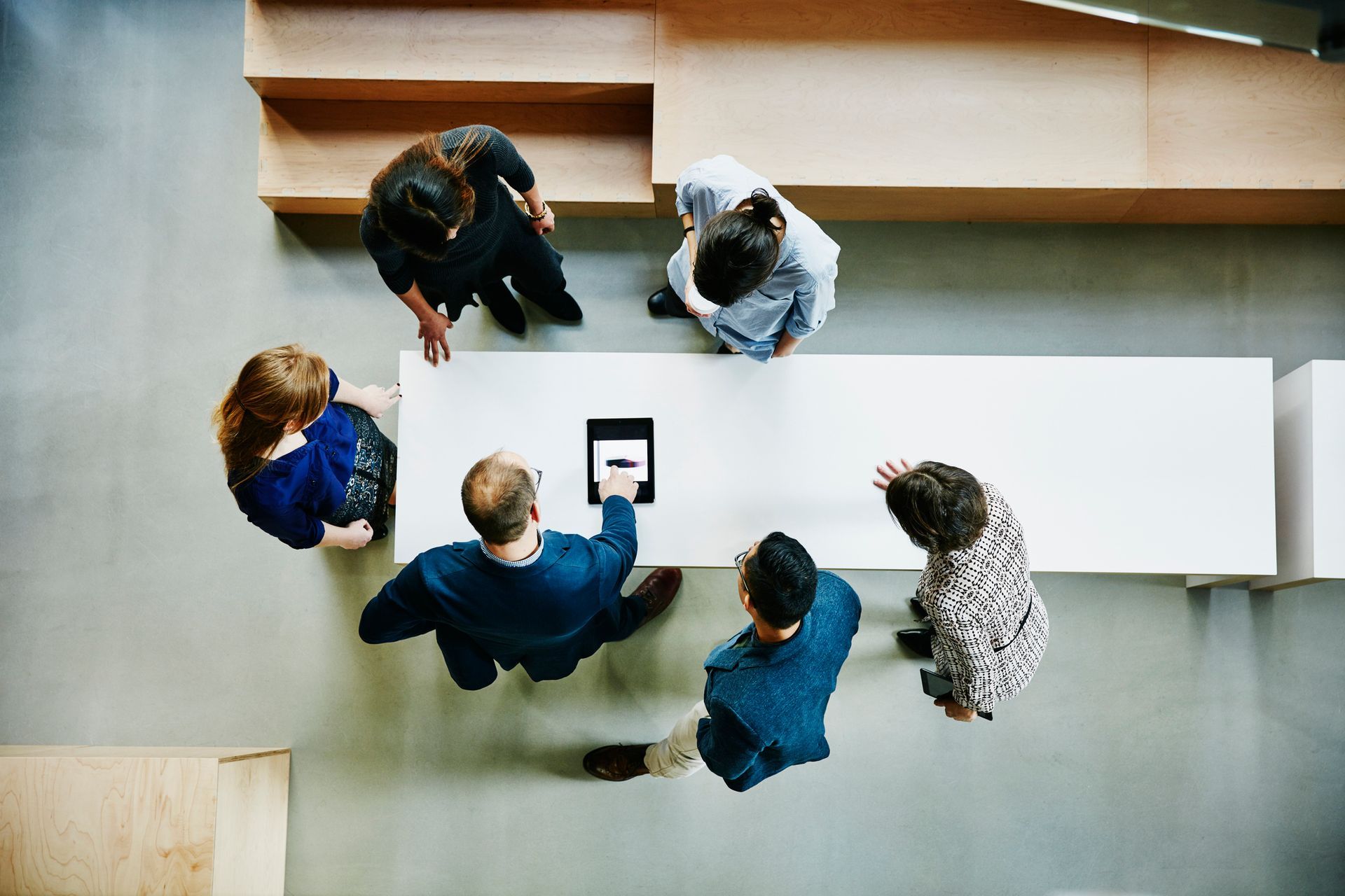 People gathered around a white table, looking at a tablet. Modern office setting with wooden accents.