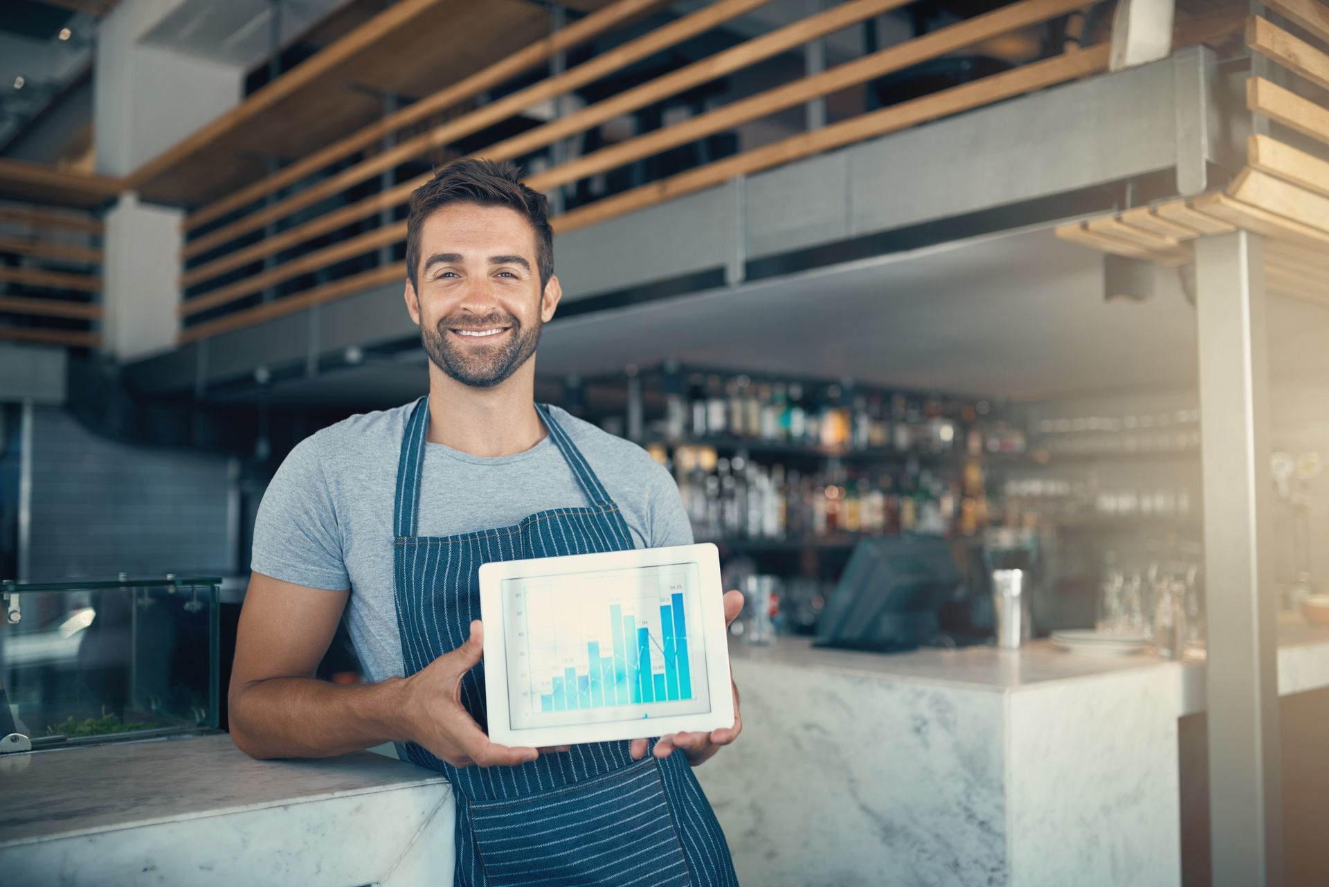 Man in apron at cafe, smiling, holding tablet with blue bar graph.