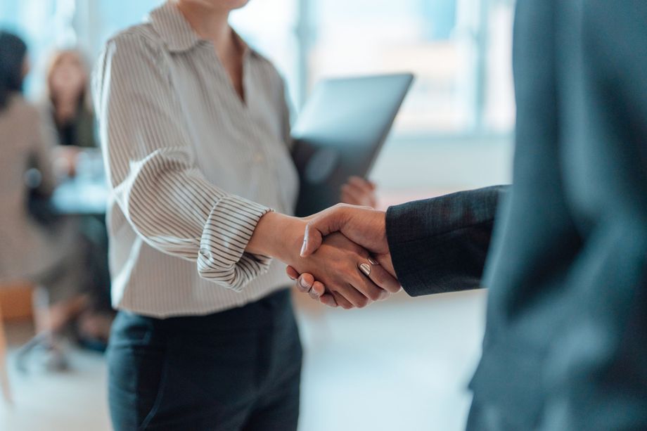 Woman in button-up shirt shakes hands with a person in a suit; office setting with others at a table.