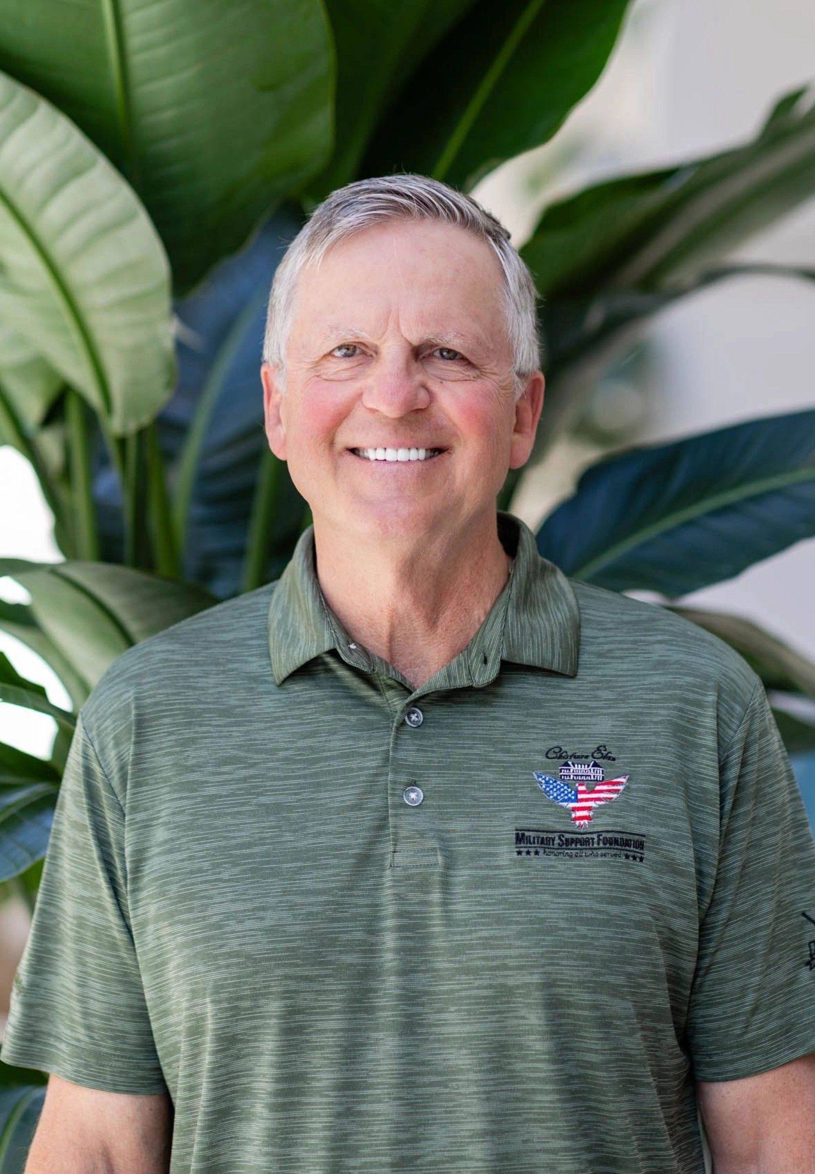 Smiling older man in a green polo shirt stands in front of large leafy plants.