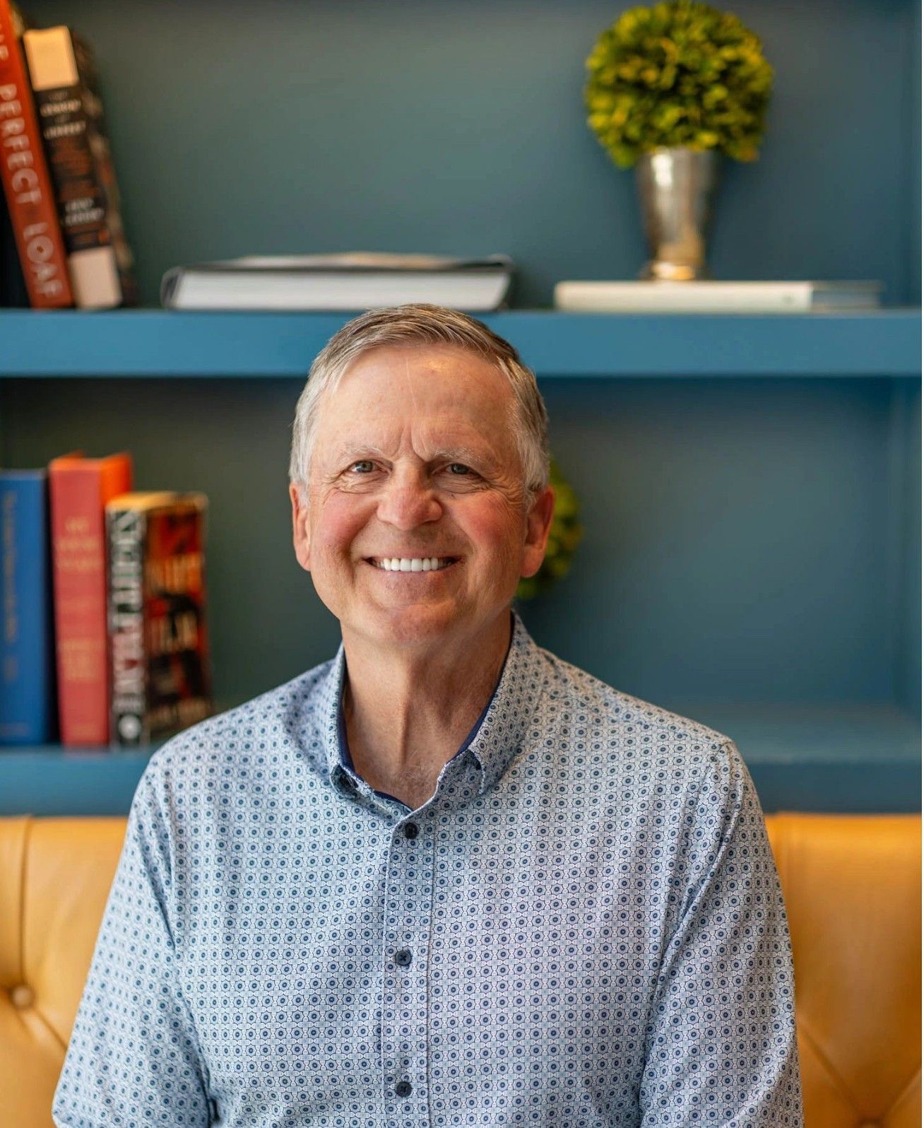 Woman in white blazer and orange pants smiles, holding a tablet, seated on a desk in an office.