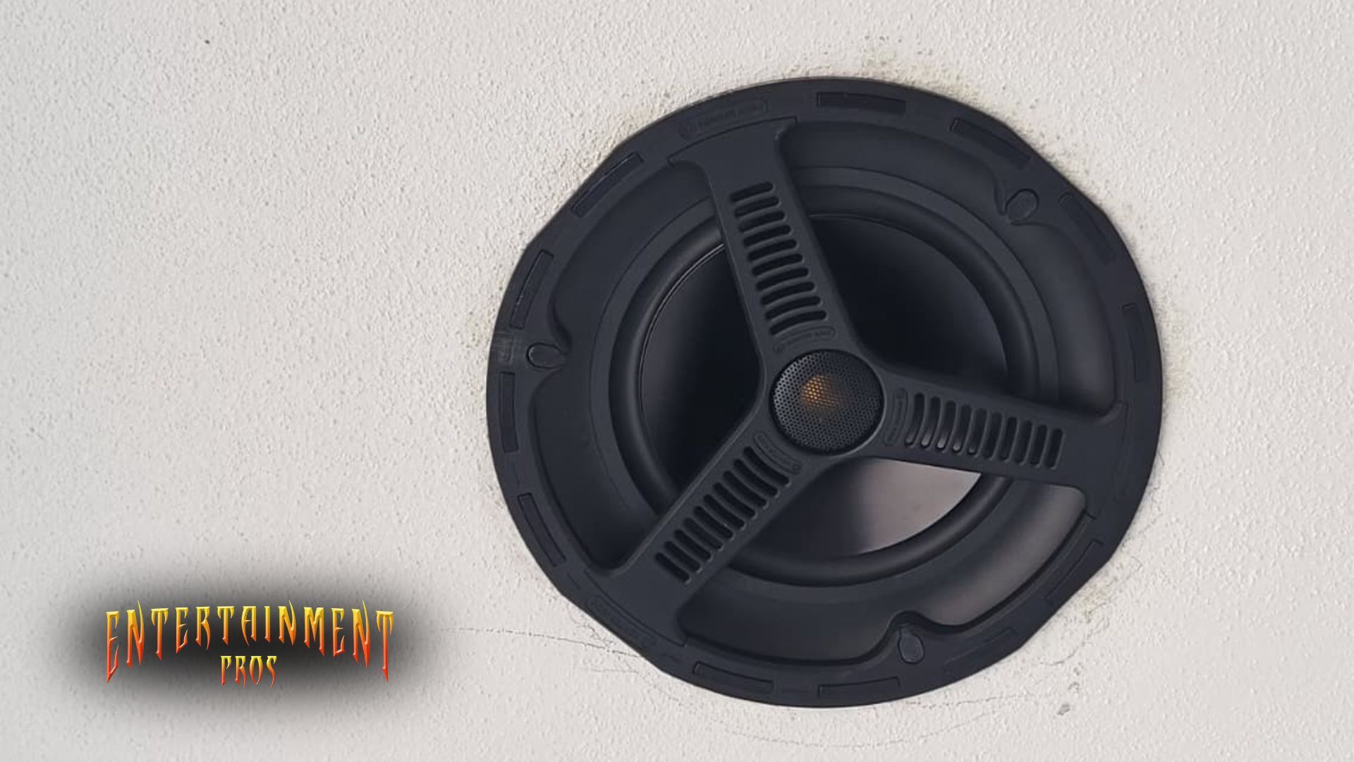 Black speaker in a white ceiling, with three radiating vent-like features.