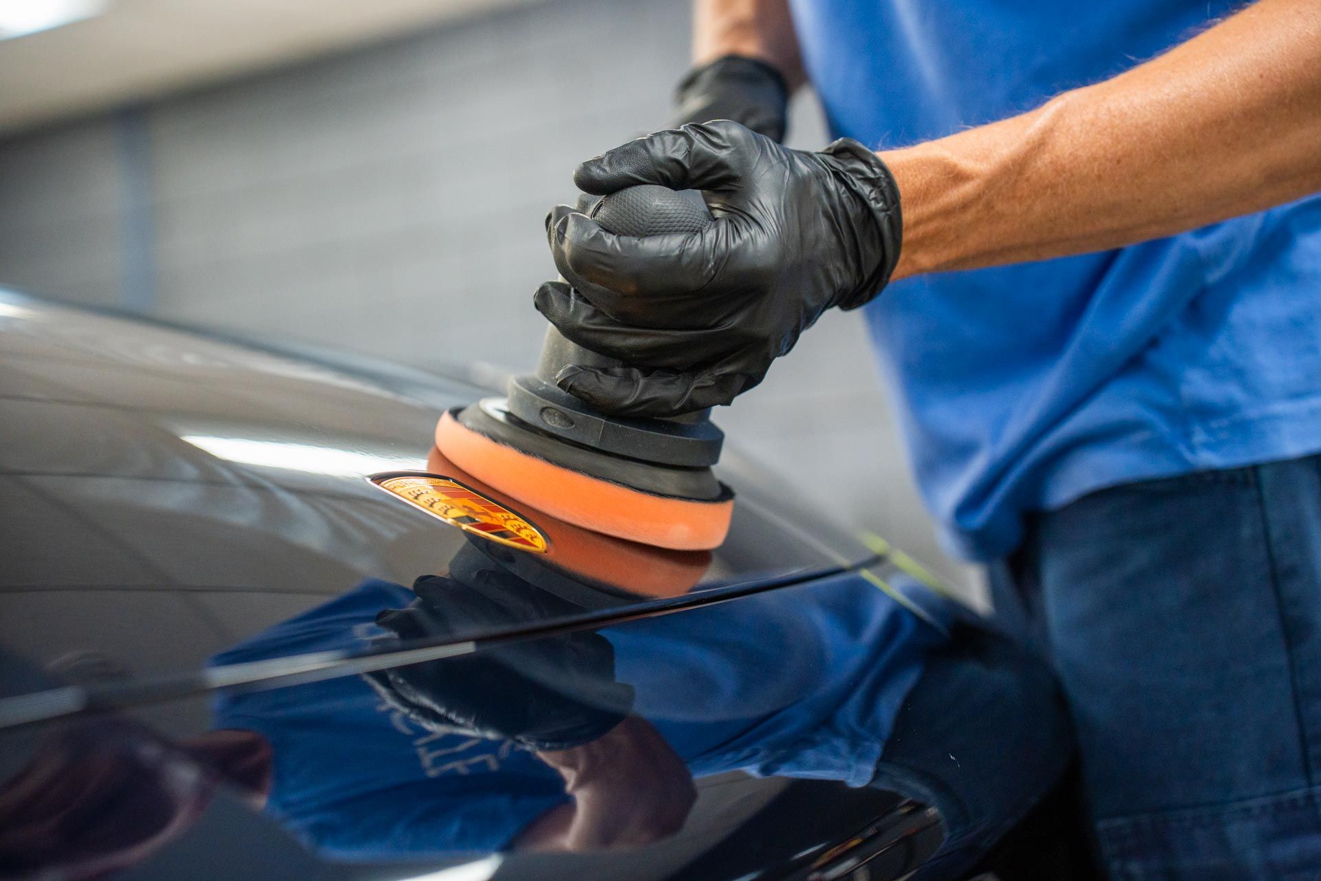 A person wearing black gloves using an orbital polisher on a car's dark surface.