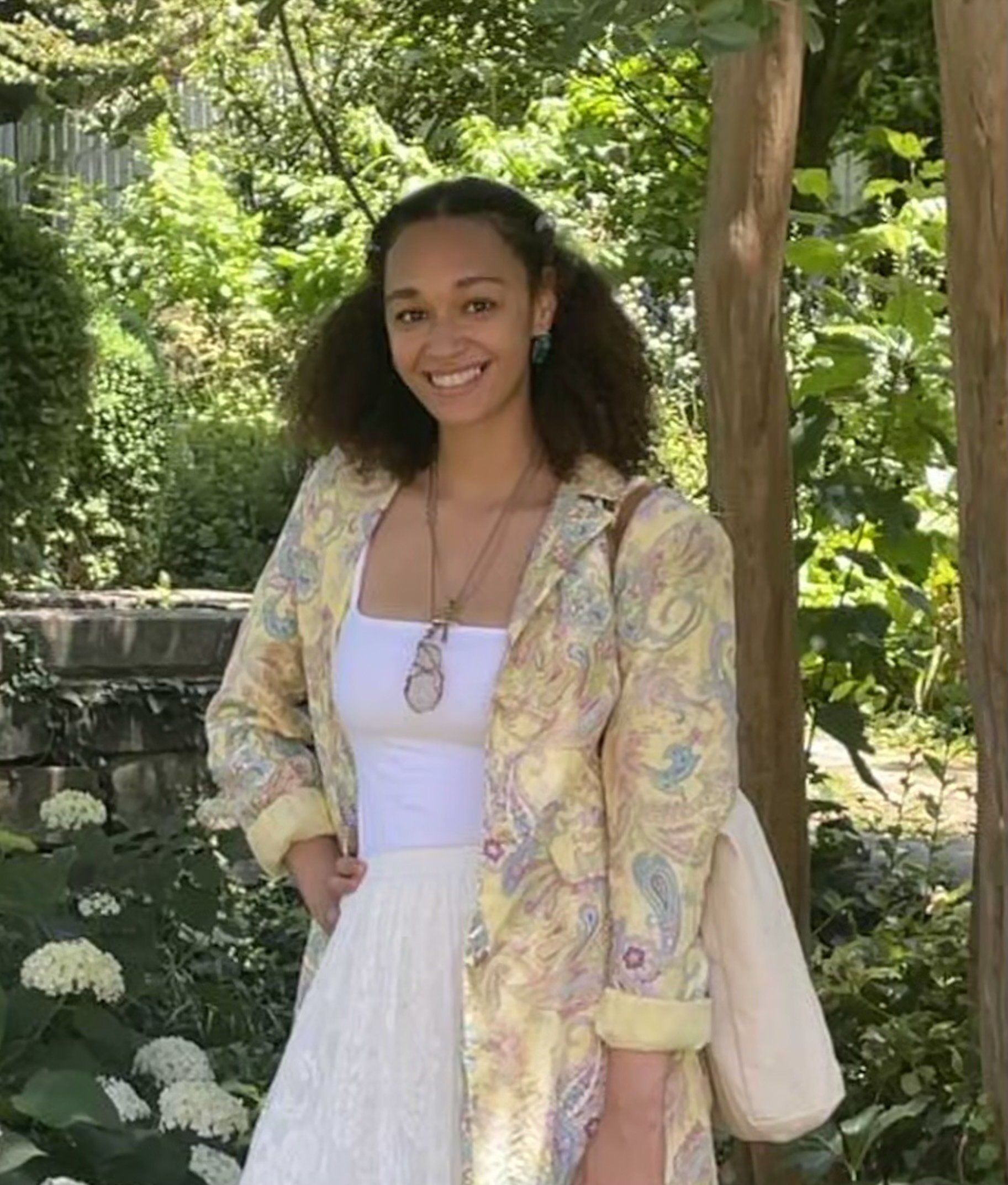 A woman in a yellow jacket and white skirt is standing in front of trees.