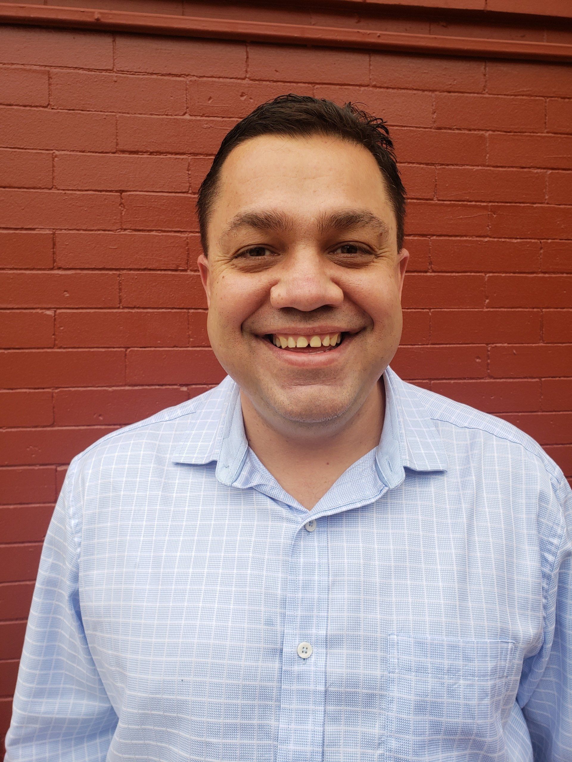 A man in a blue shirt is smiling in front of a red brick wall.
