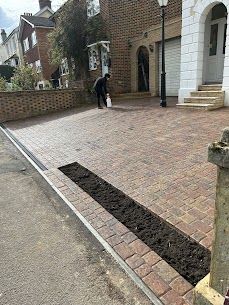 Driveway paved with brick, featuring a long, open planting bed. A person stands near the house.