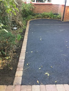 Driveway paved in black asphalt with brick edging, next to a garden of green plants and a brick house.