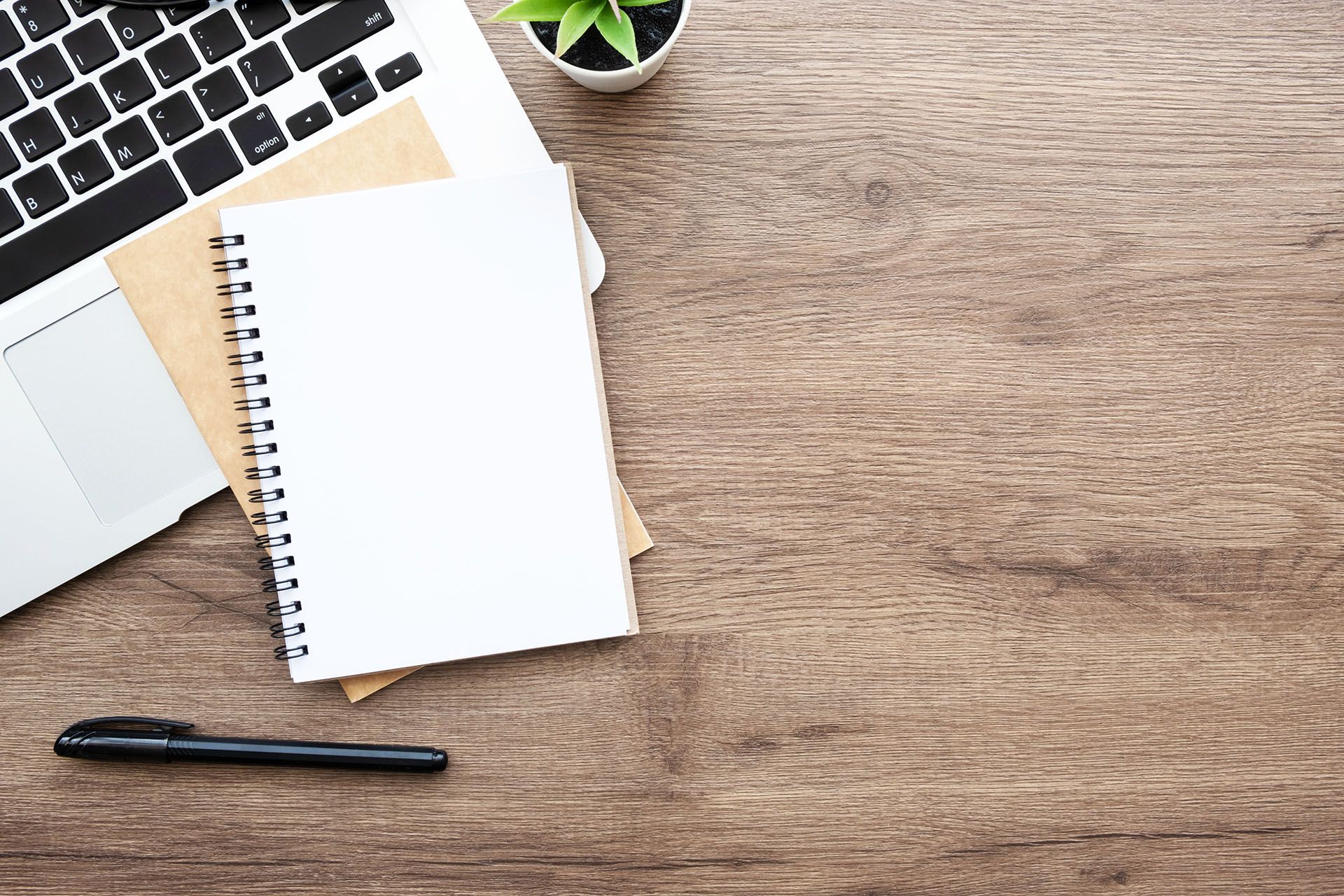 A wooden desk with a laptop , notebook , pen and plant.