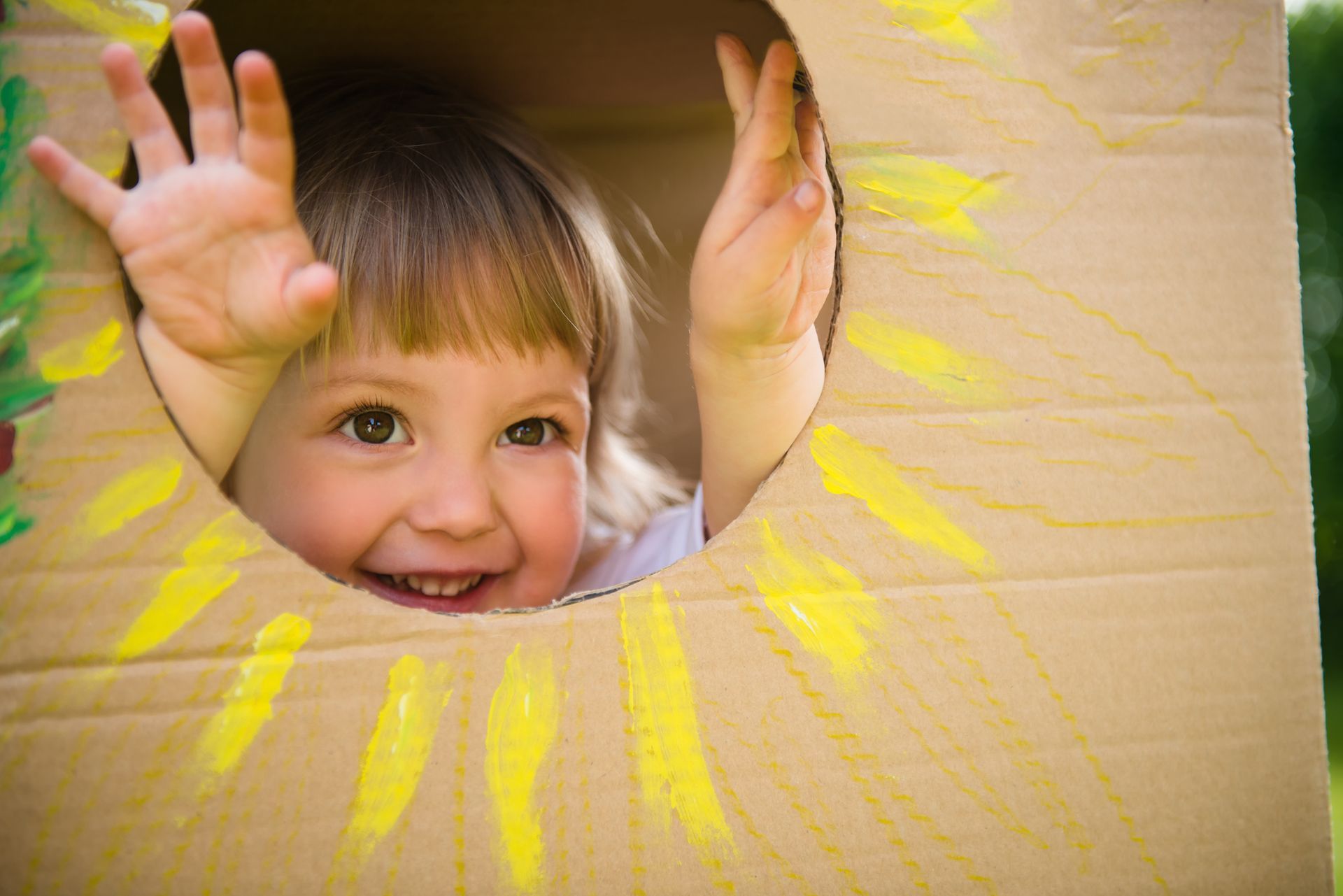 Child peeking through a hole in a box with a yellow sun painted around the hole