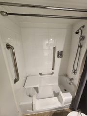 Accessible shower with grab bars and a built-in seat. White tiled walls, shower head, and curtain.