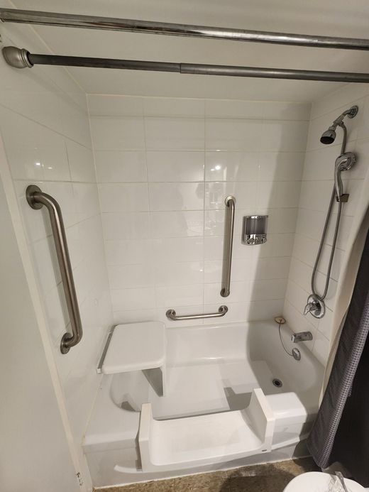 White accessible shower with grab bars and built-in seat; showerhead and curtain visible.
