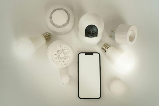 Smart home devices including lights, speakers, a camera, plug, and a phone are arranged on a white surface.