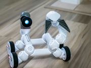 White robot with wheels and articulated arms on a wooden floor, with a blue light on its head.
