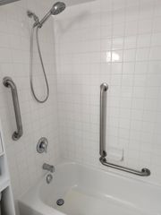 White tiled bathroom with a bathtub, showerhead, and two grab bars for accessibility.