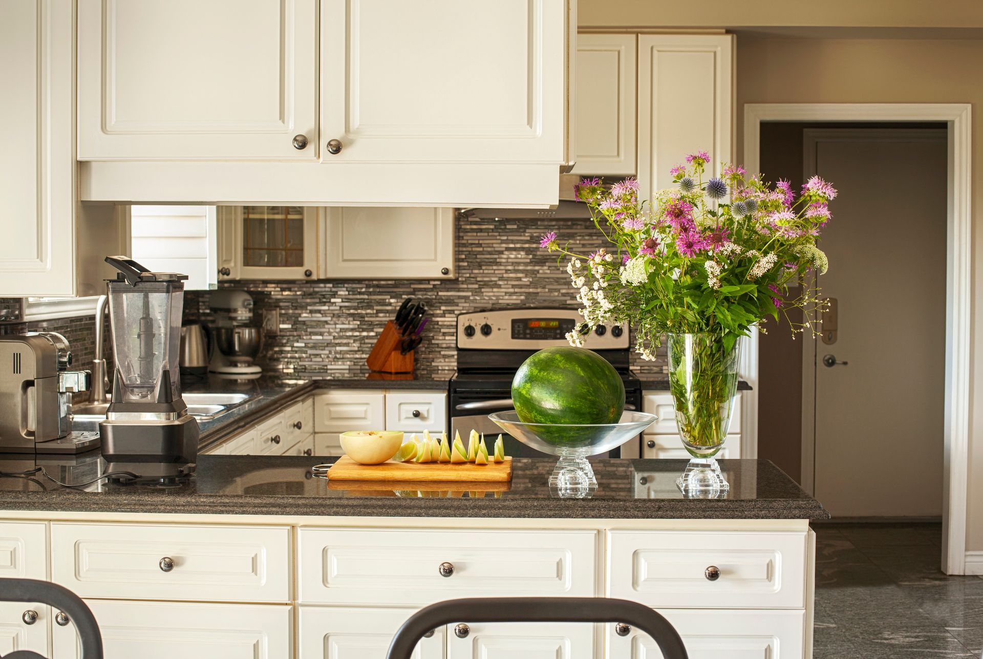 A kitchen with a watermelon on the counter and a vase of flowers.