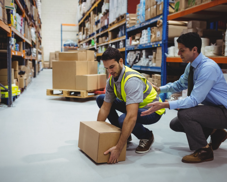 A warehouse worker, wearing a safety vest, lifts a box assisted by a man wearing business professional clothing.