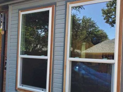 Two vertical windows with white sashes and wood frames are mounted on a gray wall; one reflects a blue sky and trees.