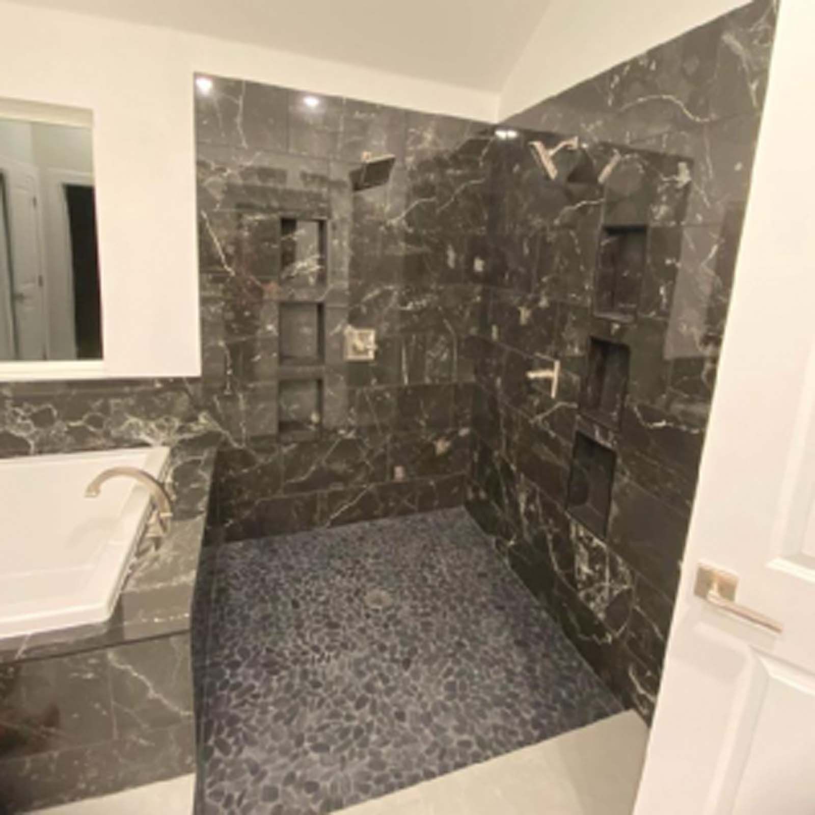 Bathroom with dark marble tile shower and stone floor, white tub.