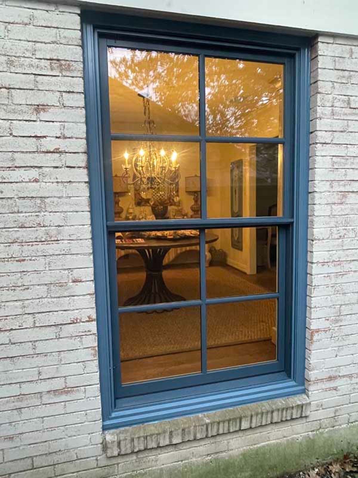 Window with dark blue frame reflects interior with chandelier and table, set against brick exterior.