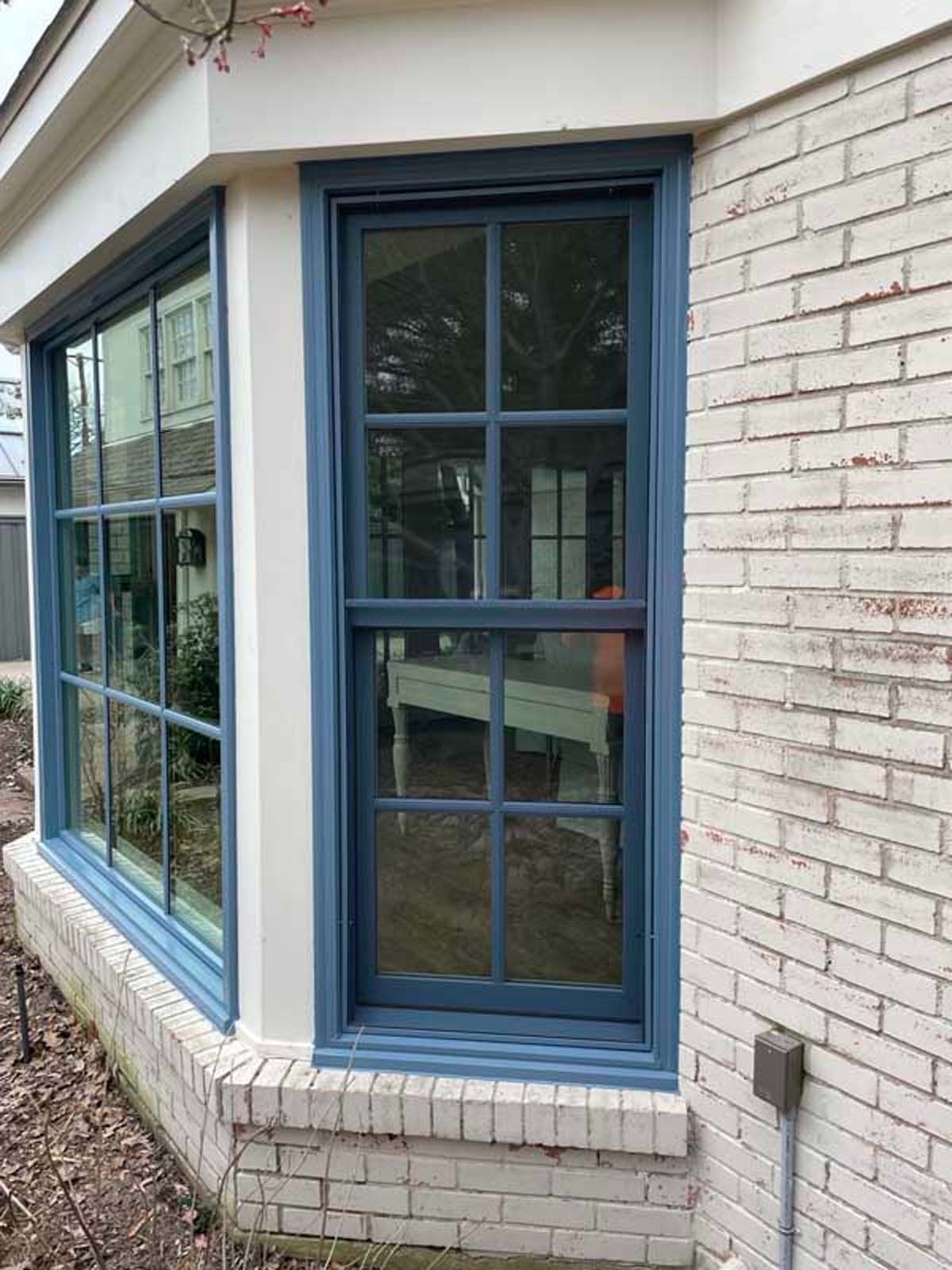 Blue-framed windows on a brick house corner, with white trim and a small bush in front.