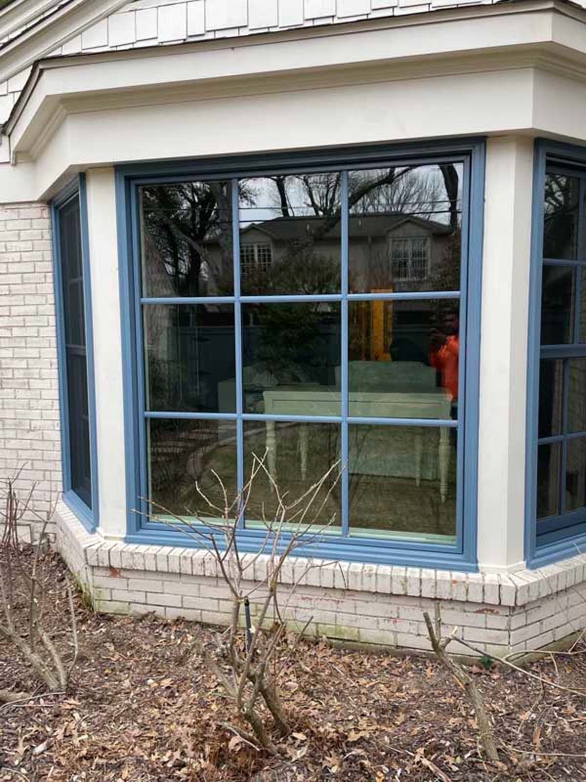 Bay window with blue trim, brick foundation, reflecting trees and a house.
