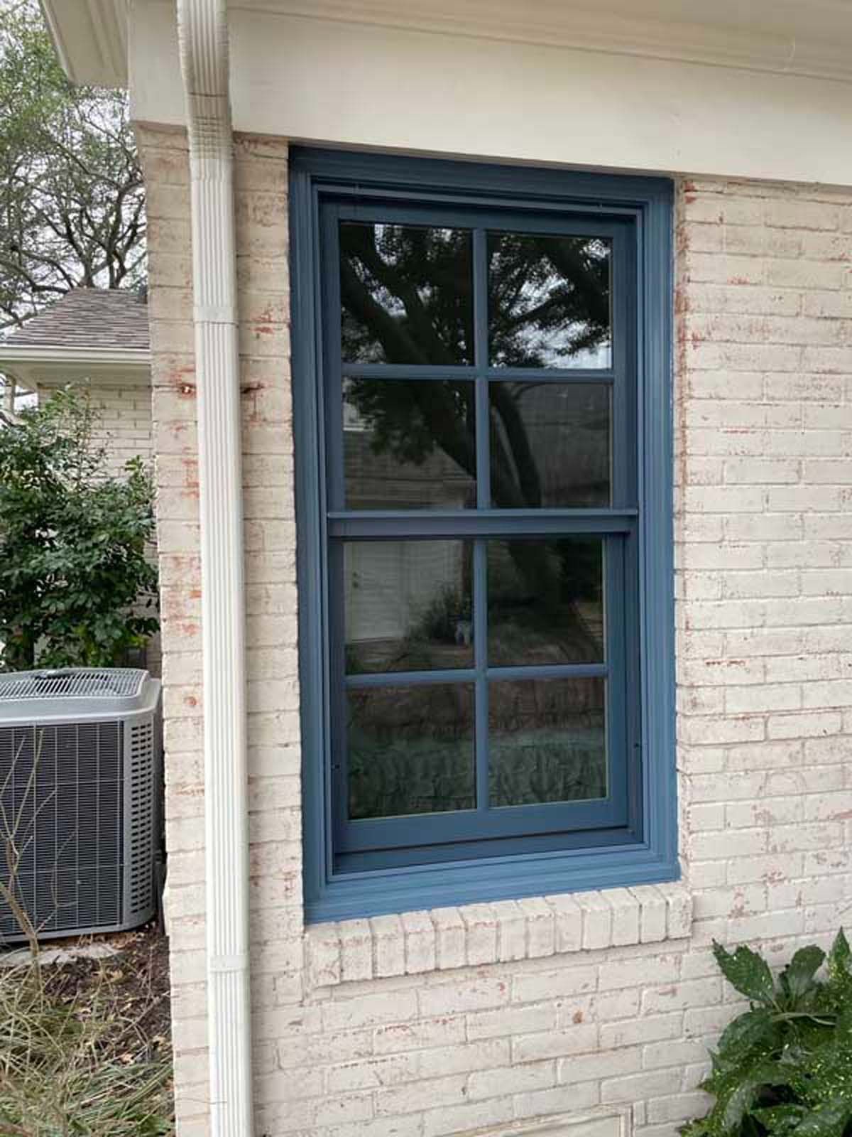 Blue-framed window on a brick building with a reflection of trees, and an air conditioning unit.