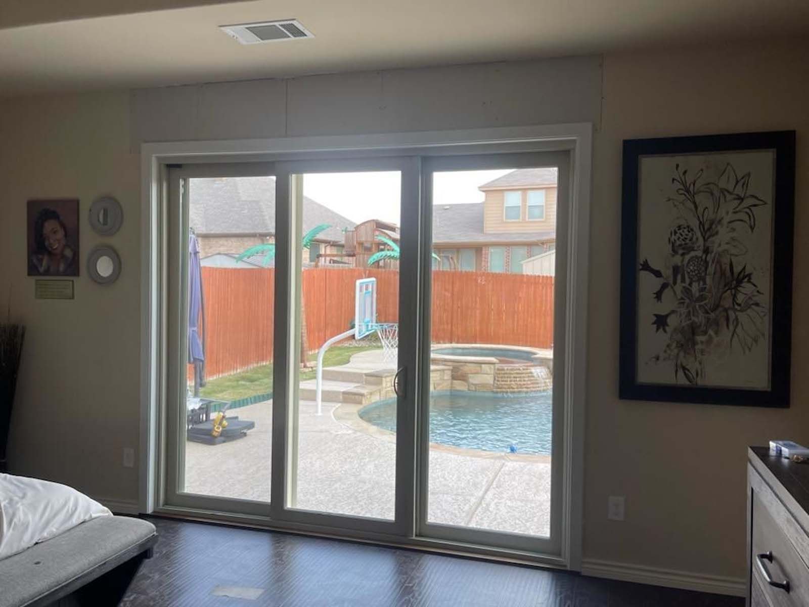 Sliding glass doors open to a backyard with a pool. A brown fence surrounds a house, and a wall art piece hangs on the right.