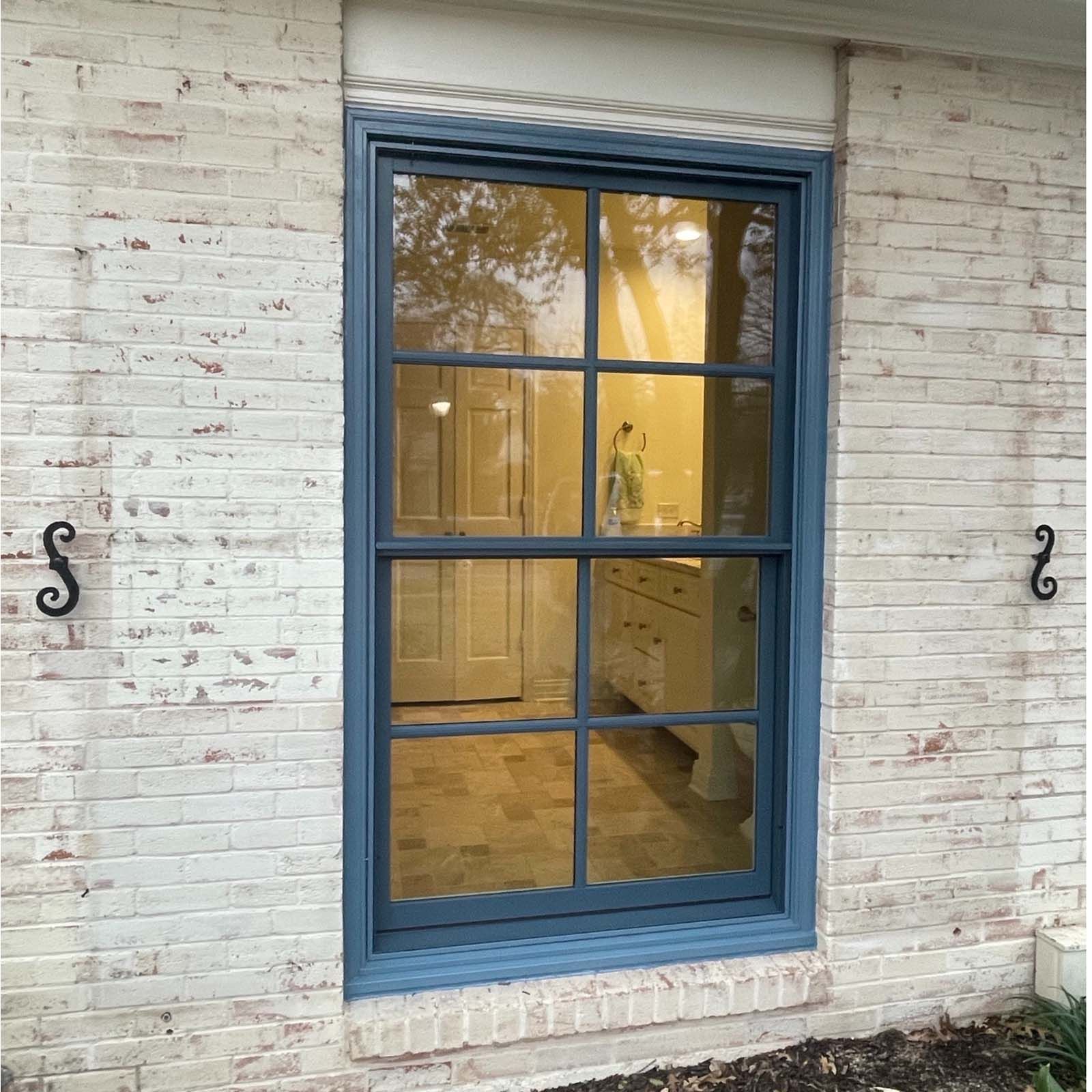 Blue-framed window in brick wall, reflecting interior with cabinets and a door, decorative ironwork on either side.