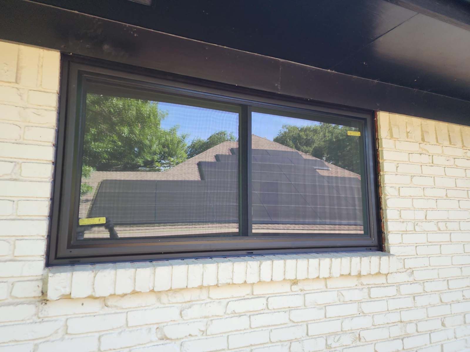 Dark-framed sliding window on a white brick exterior, reflecting trees and a house under a blue sky.