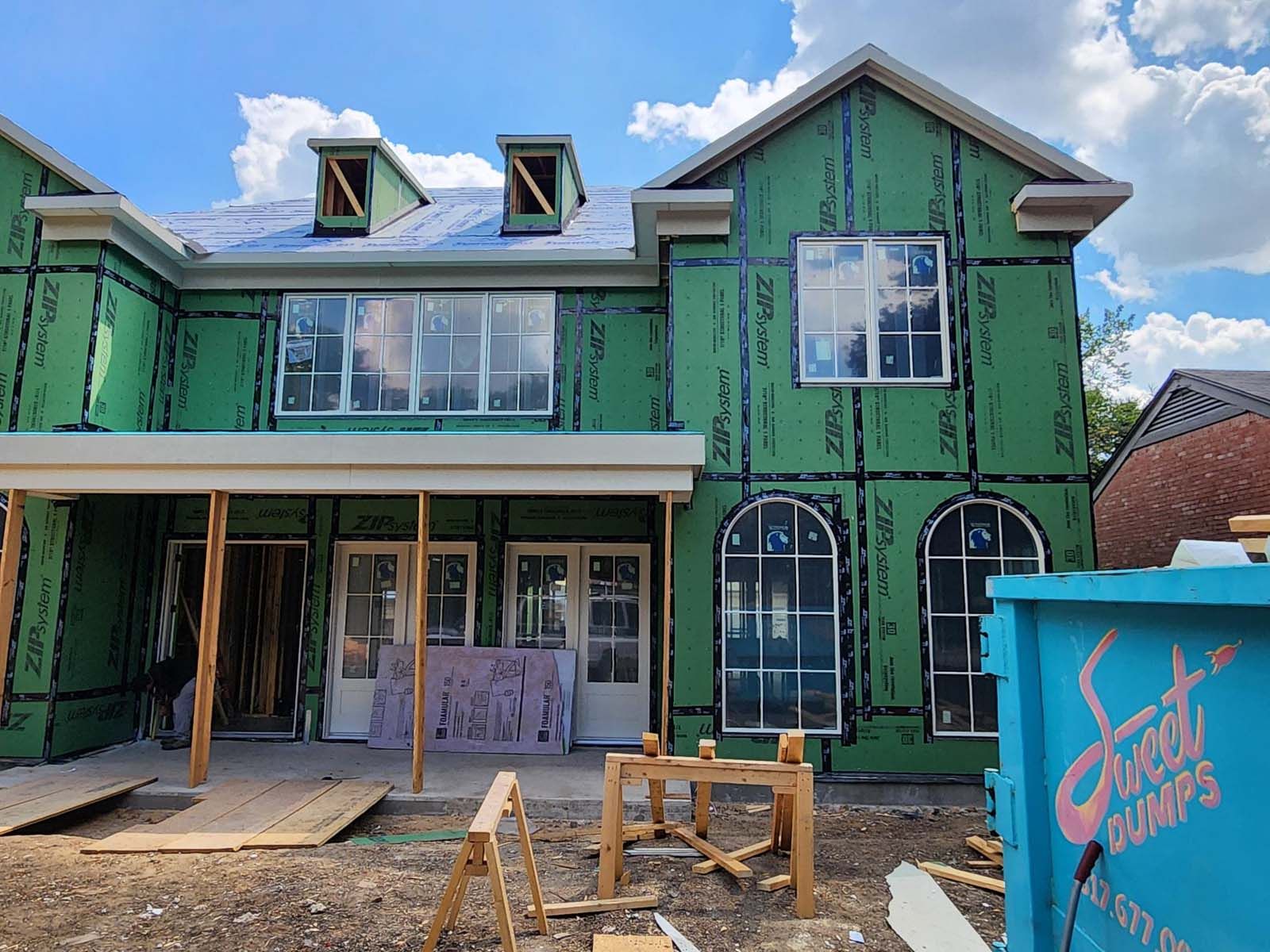 House under construction, green sheathing, white windows, blue dumpster, bright sunny day.