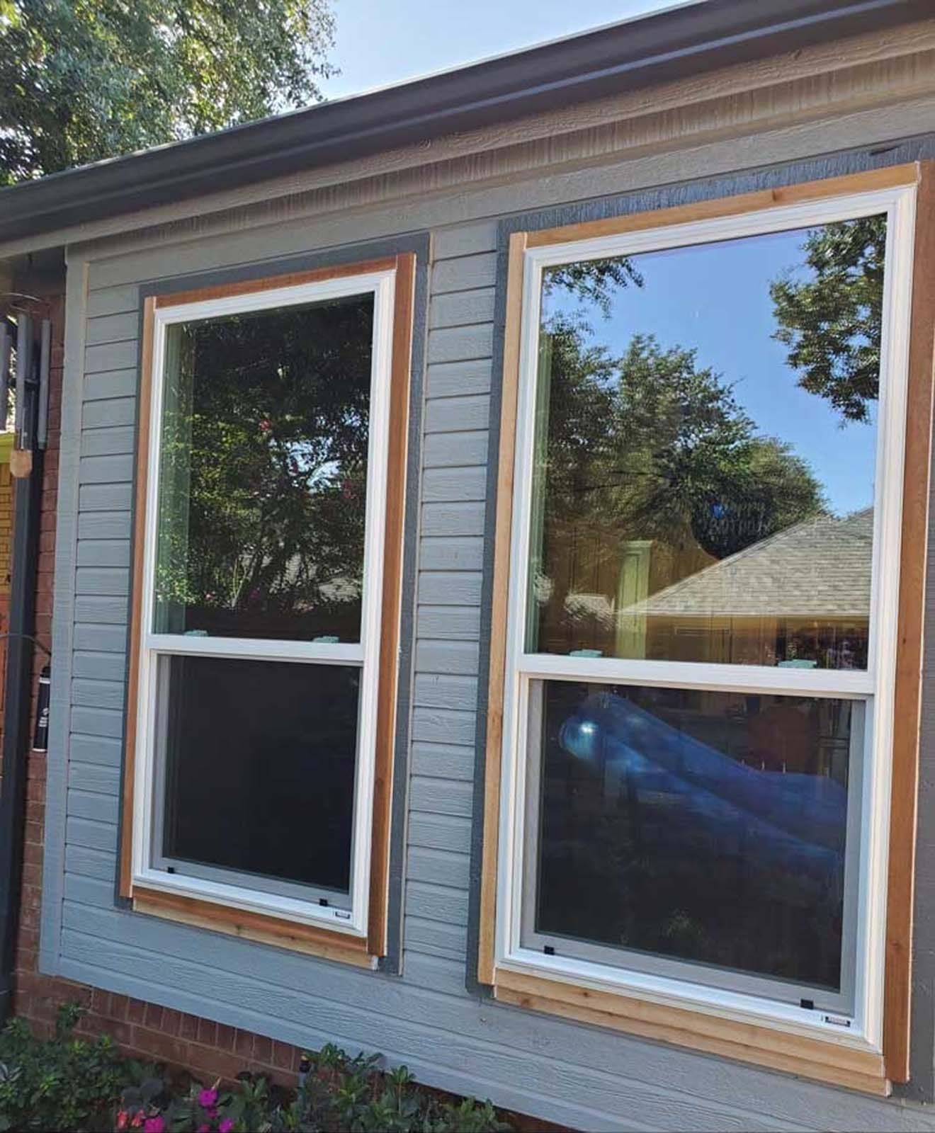 Two newly installed windows with wood frames and white trim on a blue-gray siding wall, reflecting the sky.