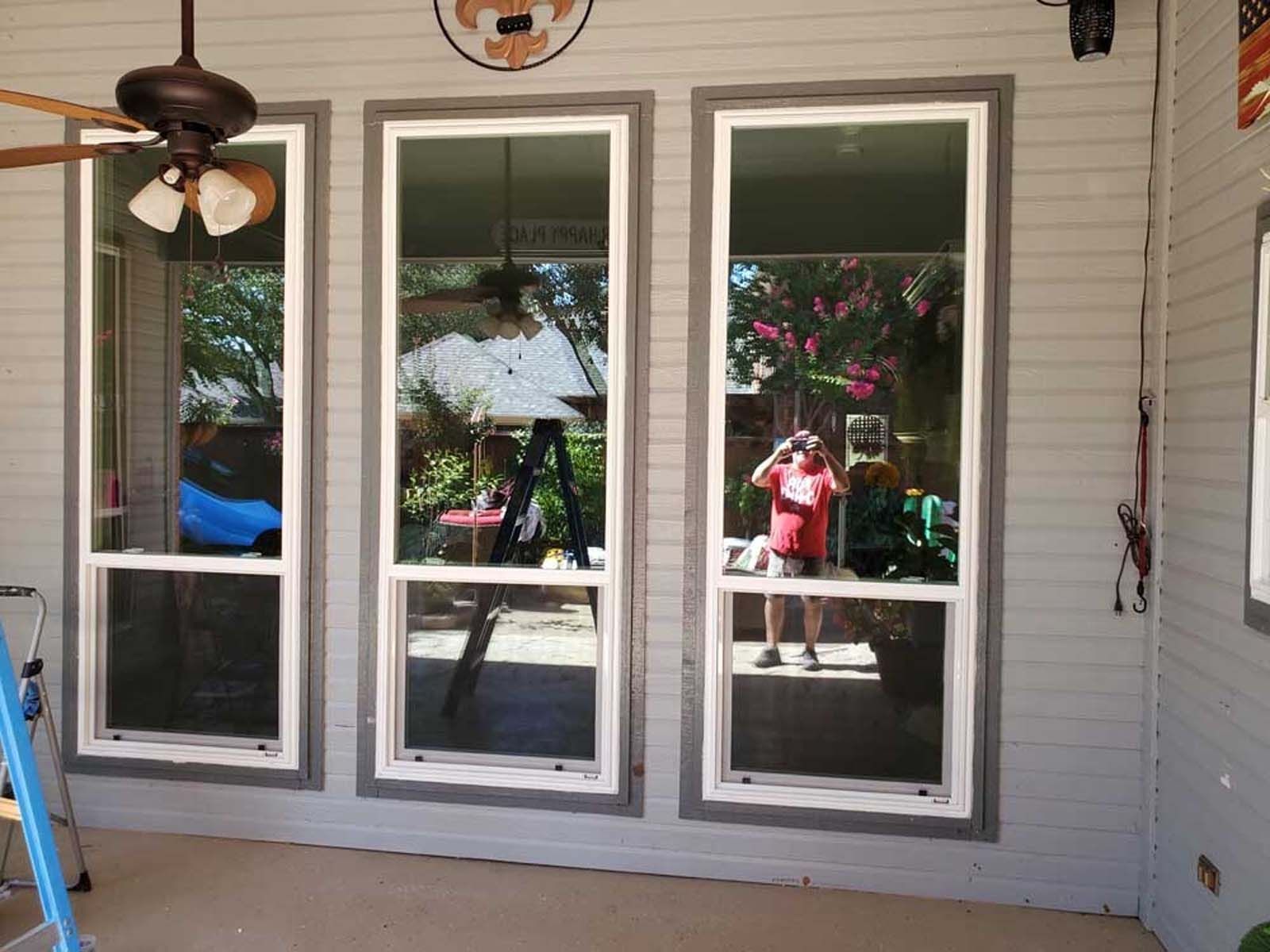 Three tall windows on a gray-sided porch, reflecting a person, trees, and a ladder.