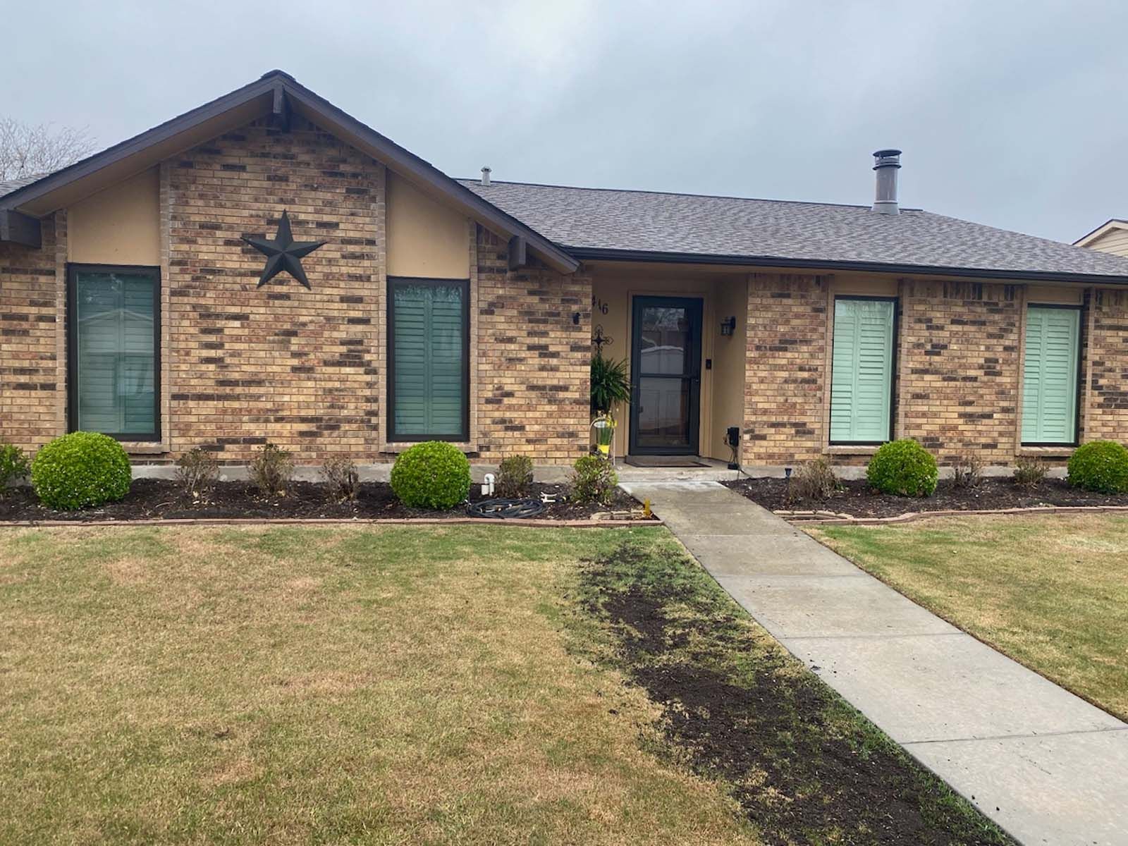 Single-story brick house with brown roof and star, sidewalk, and patchy lawn.