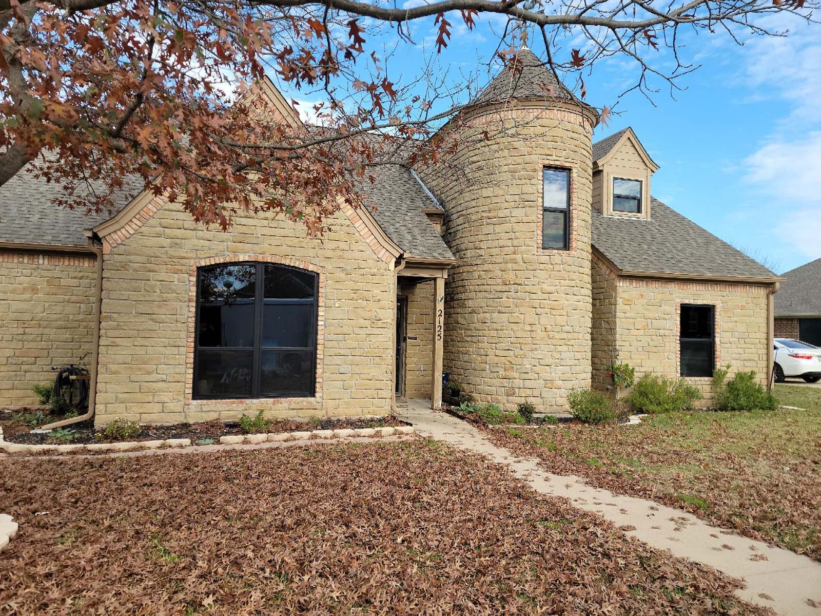 Tan brick house with turret, arched window, and walkway. Brown leaves on ground, blue sky.